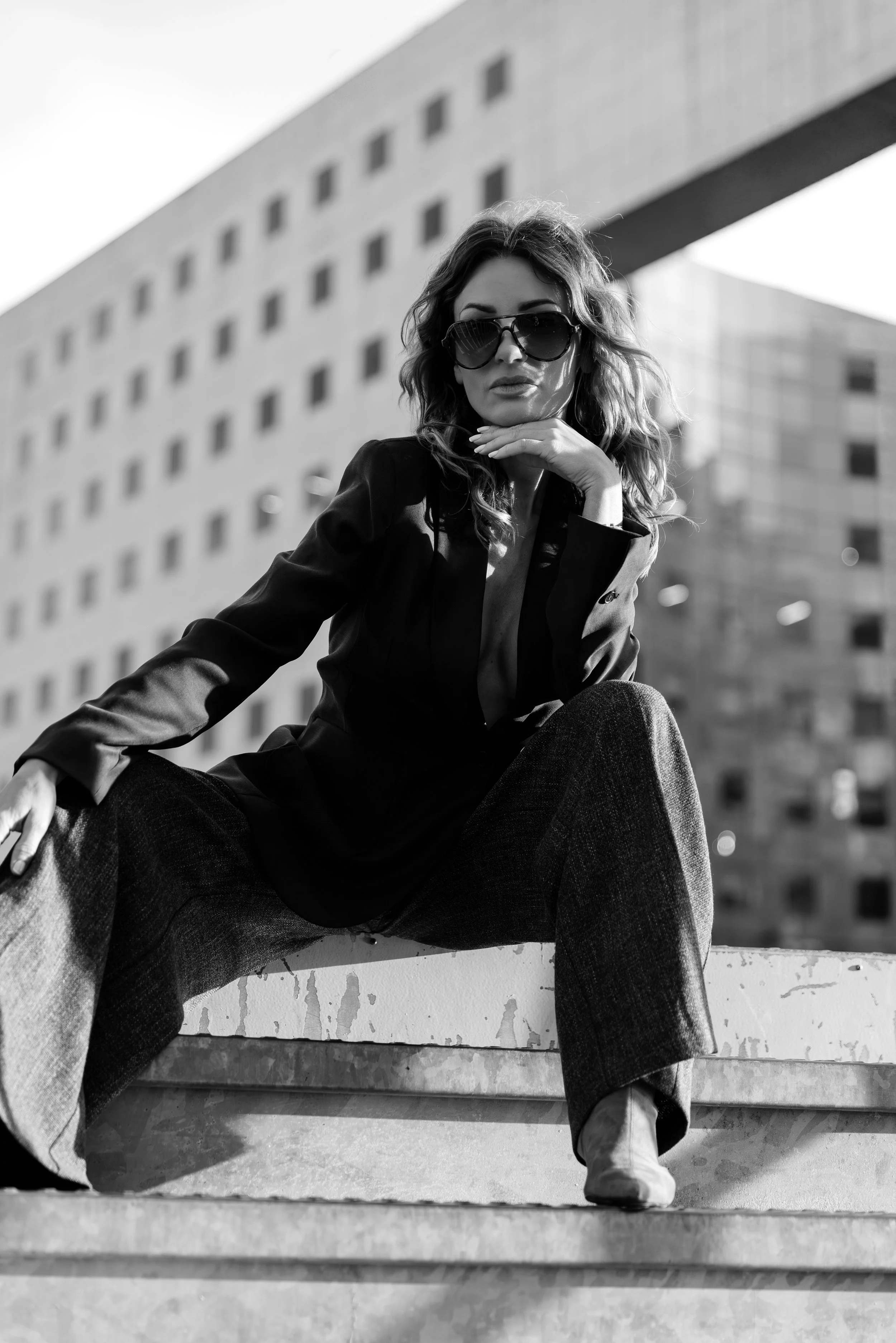 Une femme posant en extérieur, portant des lunettes de soleil et une tenue sombre, avec un bâtiment moderne en arrière-plan.