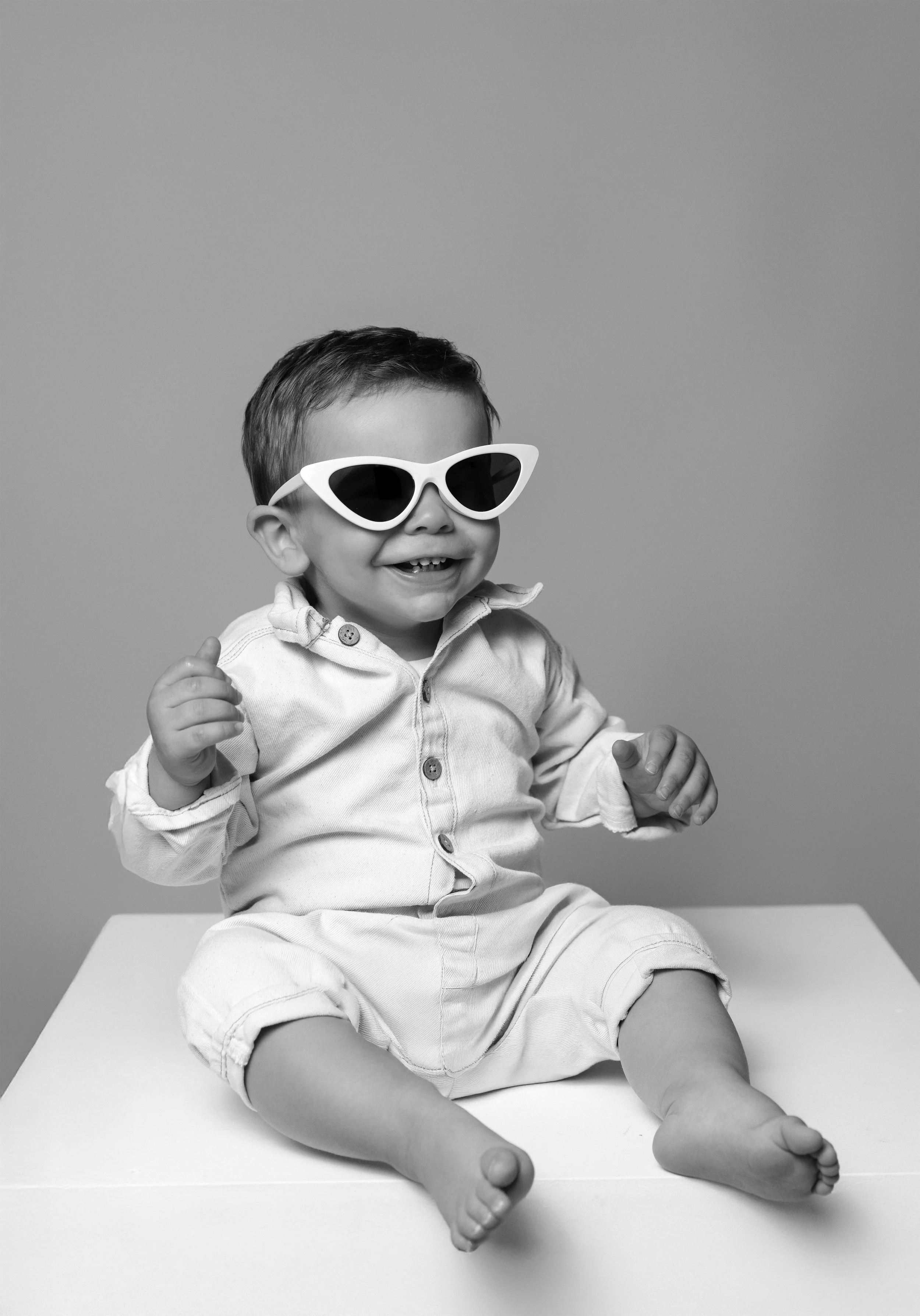 Un bébé souriant portant des lunettes de soleil blanches en forme d'ailes, assis sur une surface blanche, en noir et blanc.