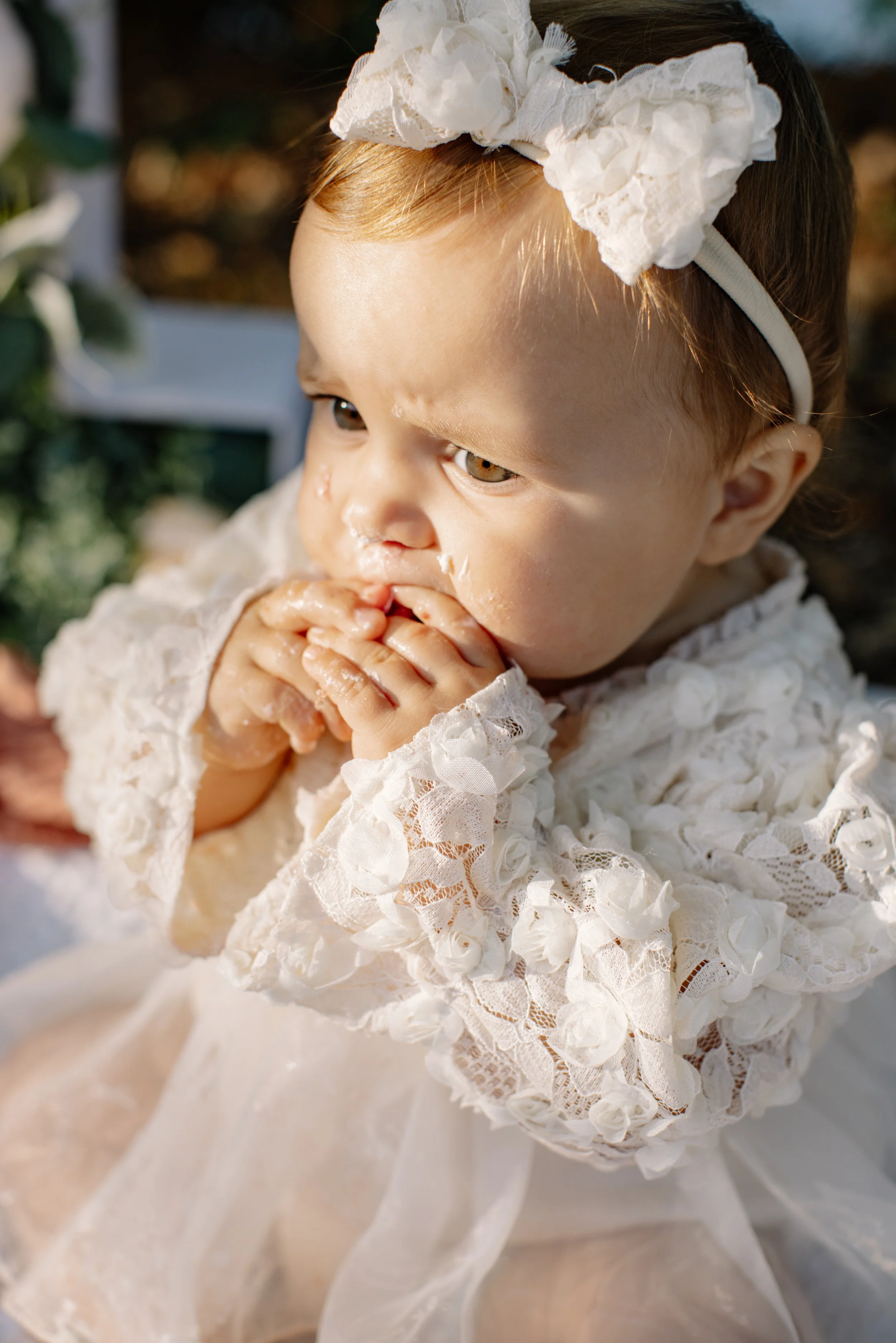 Jeune enfant en robe de dentelle blanche, portant un bandeau blanc avec un grand nœud en tissu, mangeant une pâtisserie, vue de face, lumière naturelle, décor flou en arrière-plan.