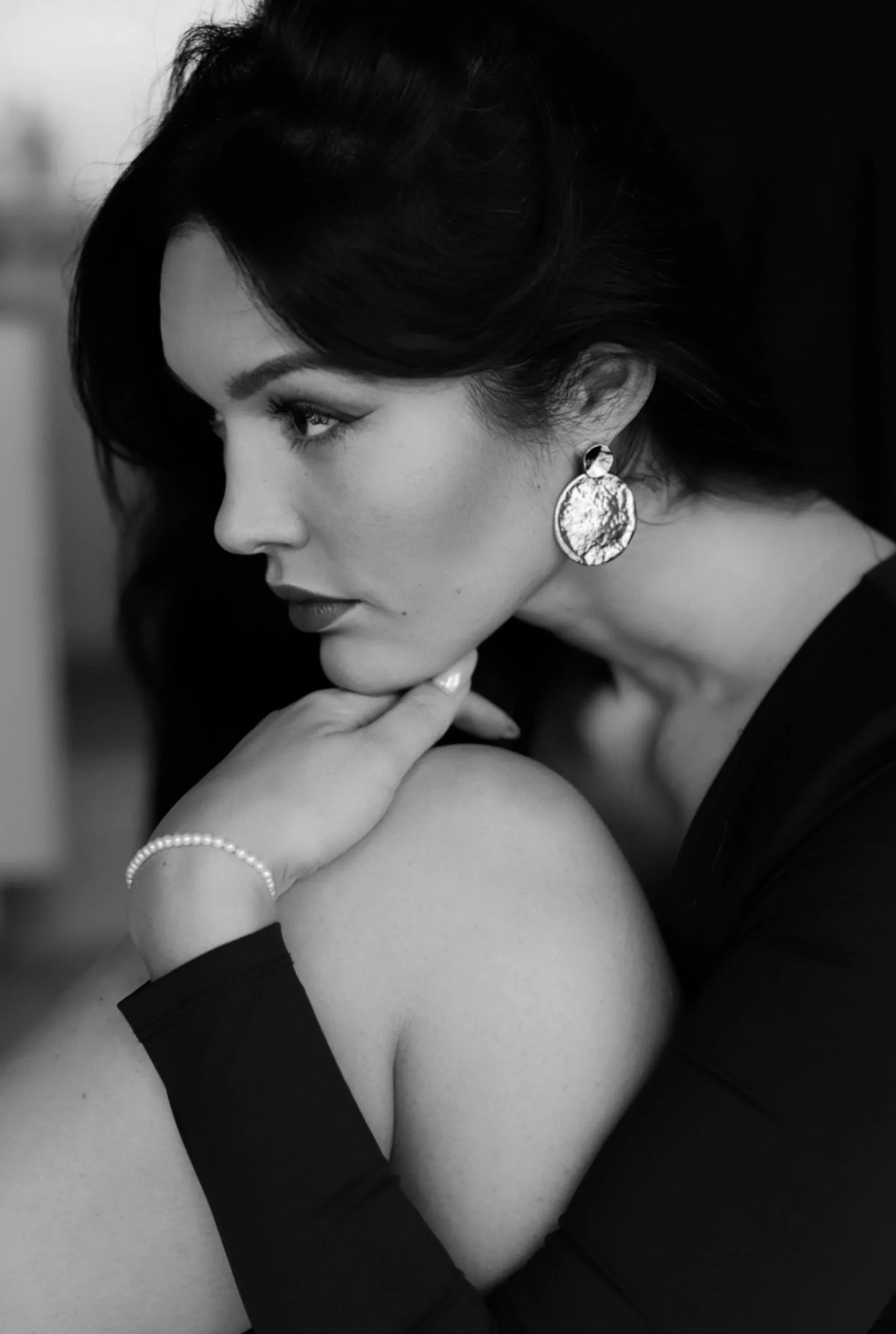 Portrait en noir et blanc d'une femme élégante regardant sur le côté, portant une grande boucle d'oreille en pierre et une bracelet de perles, avec les cheveux courts et ondulés.