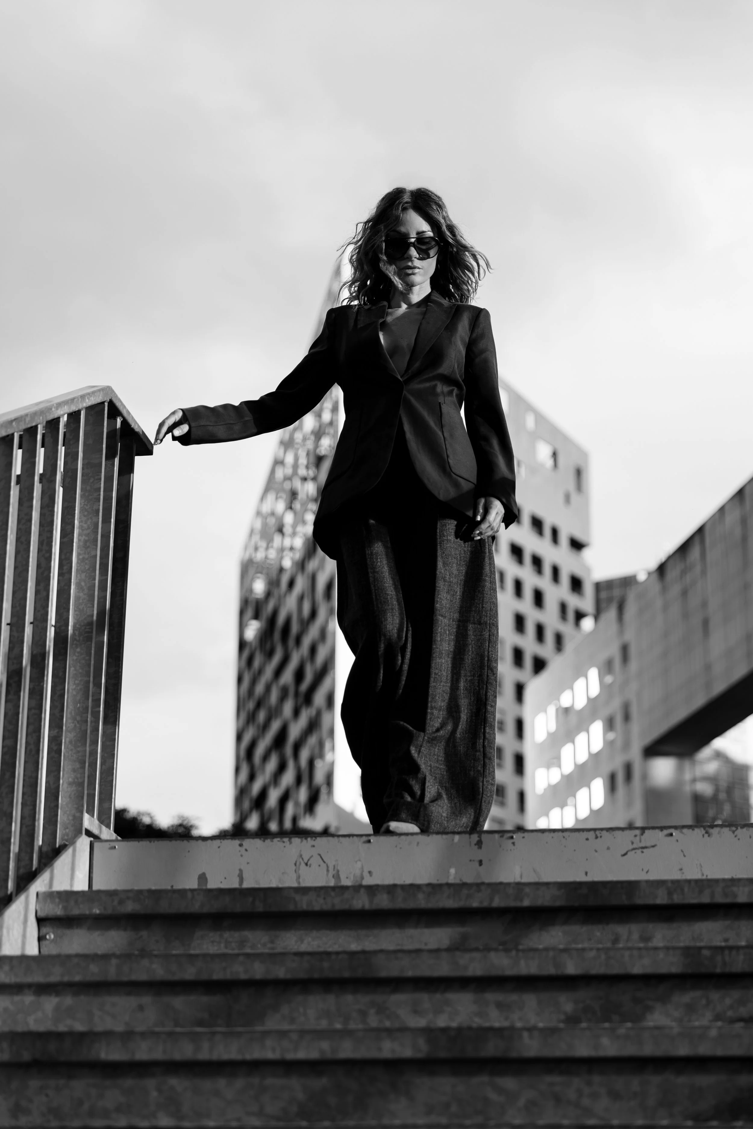 Femme portant un costume sombre descend un escalier en ville, bâtiment moderne en arrière-plan, en noir et blanc.