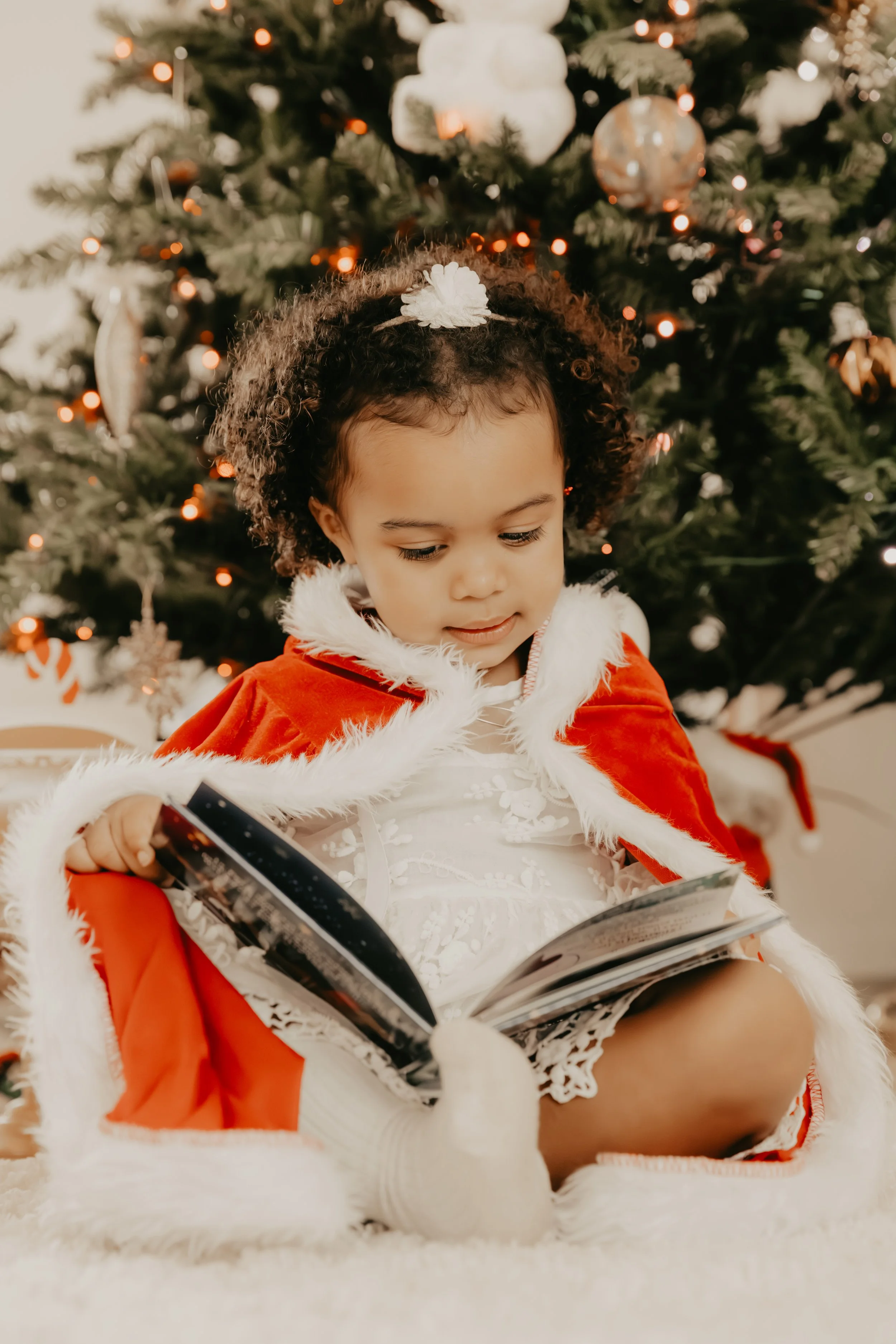 Jeune fille en tenue de fête avec cape rouge bordée de fourrure blanche, assise par terre, lisant un livre devant un sapin de Noël décoré.