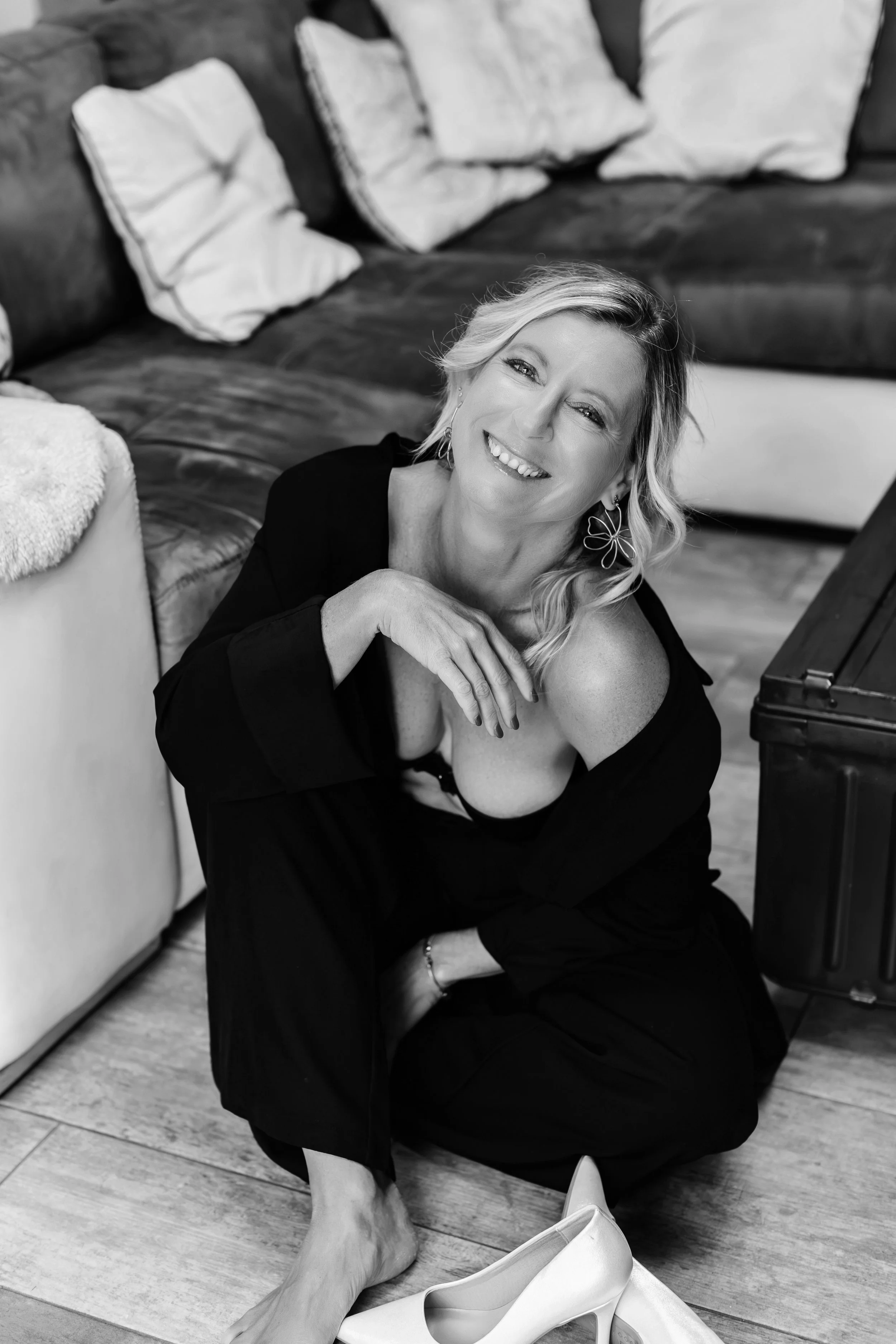 Une femme souriante, assise au sol, dans un intérieur, avec des coussins sur un canapé en arrière-plan, des chaussures blanches devant elle, une mallette à côté.