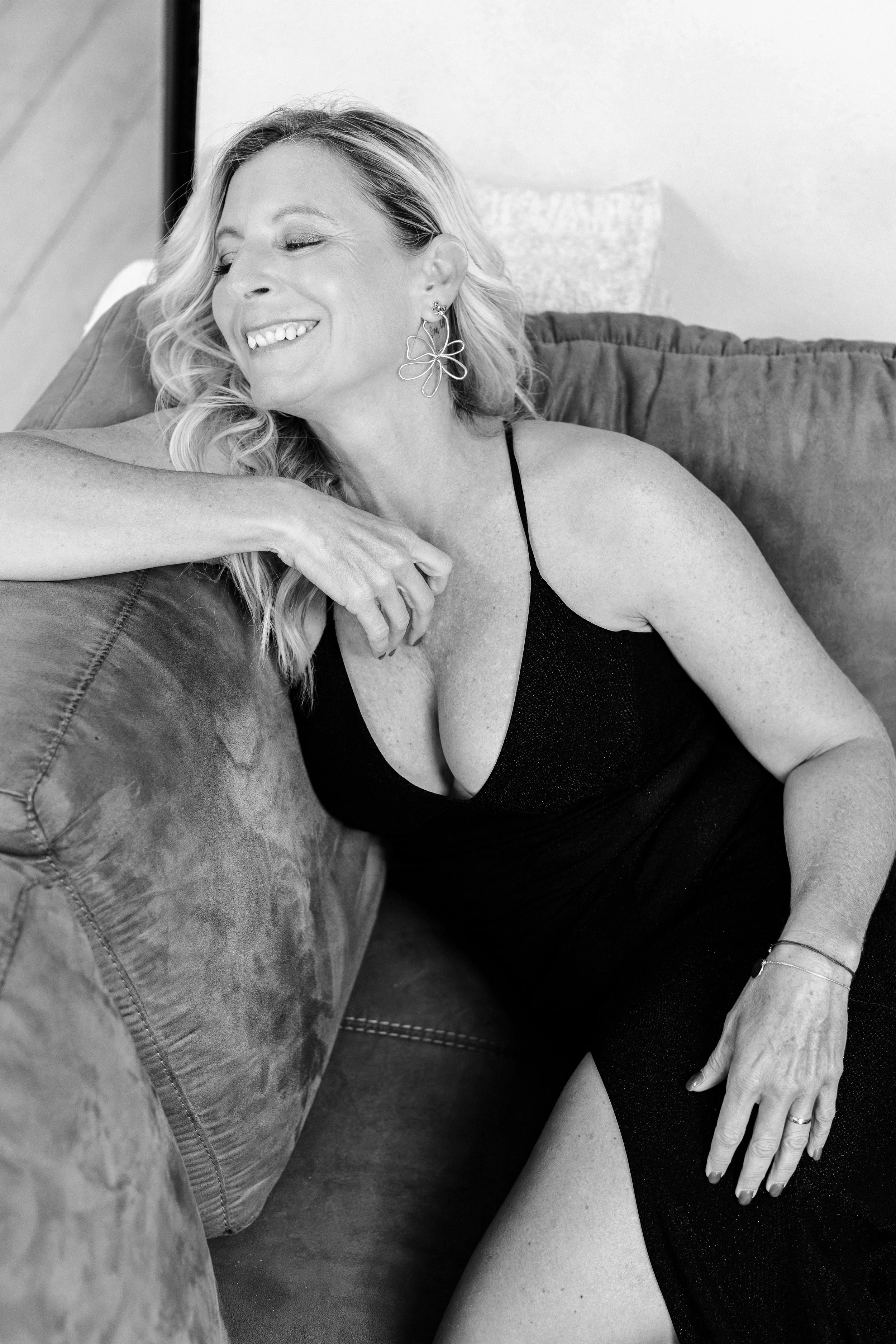 Une femme souriante, aux cheveux ondulés, portant un déguisement noir avec des bretelles, assise sur un canapé en velours, avec des boucles d'oreilles en forme de fleur et un bracelet bracelet