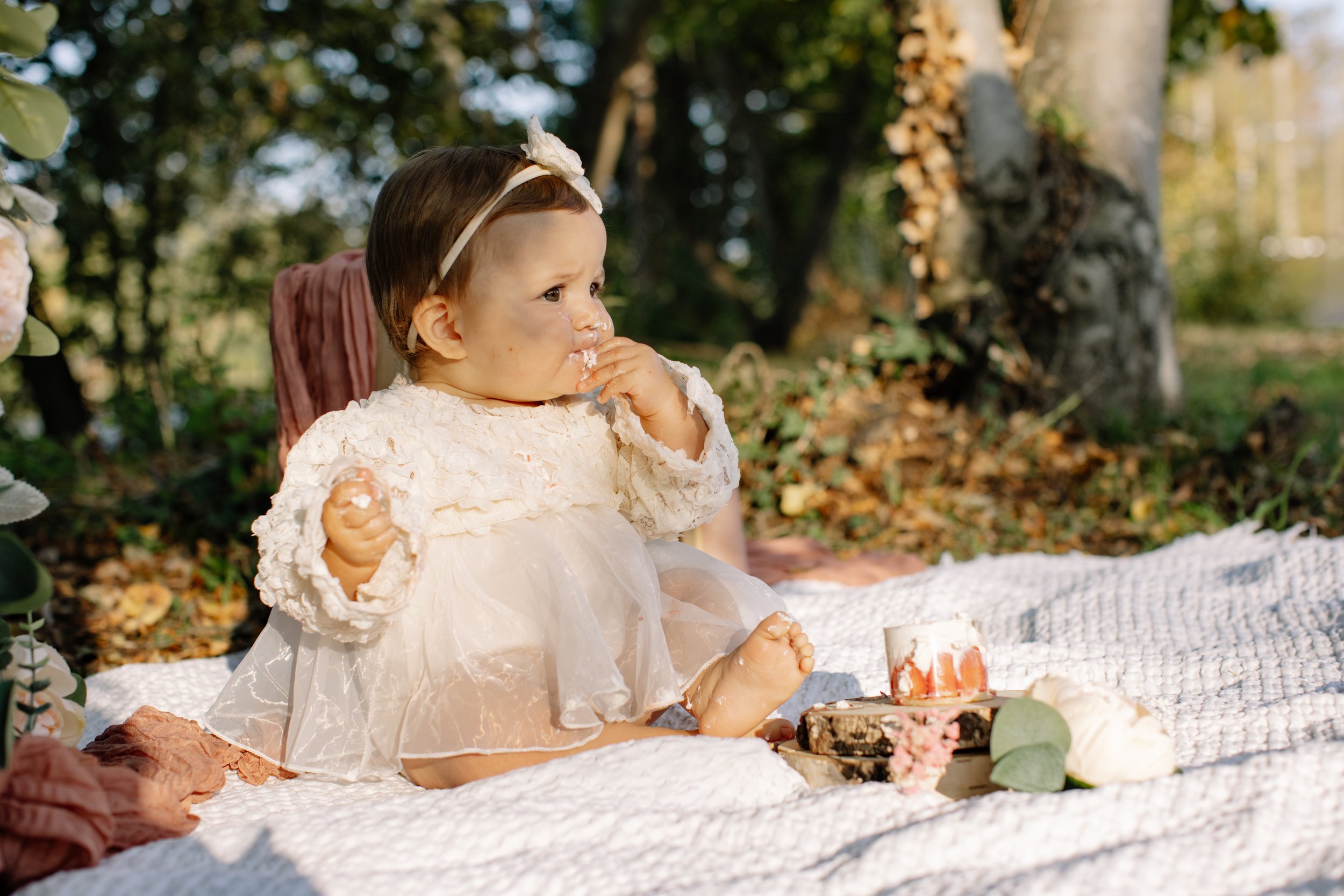 Un bébé assis sur une couverture lors d'un pique-nique, avec un gâteau et des fleurs dans un environnement forestier.