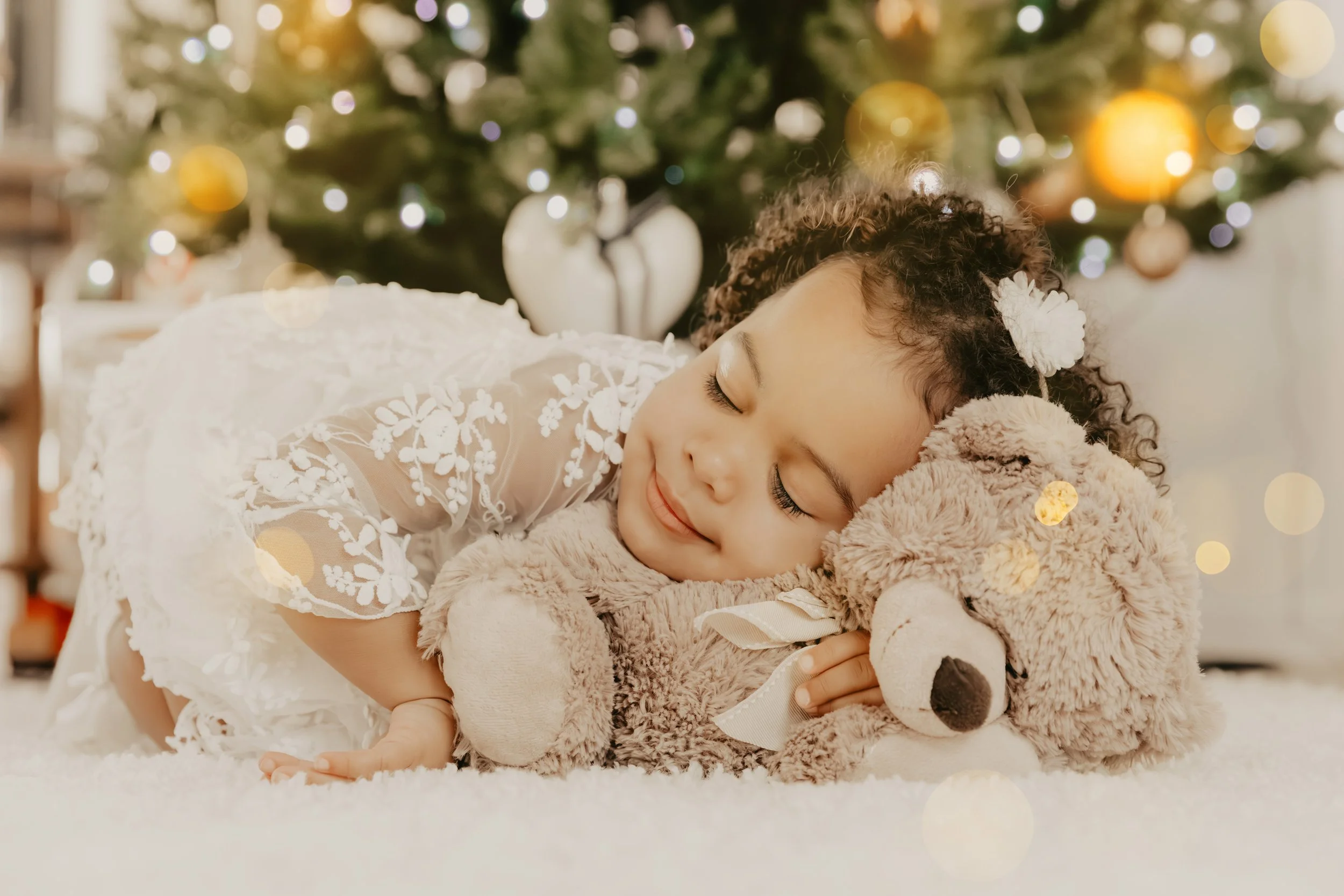 Une jeune fille joyeuse avec des cheveux bouclés, portant une robe blanche en dentelle, dort en câlinant un ours en peluche marron devant un sapin de Noël décoré.