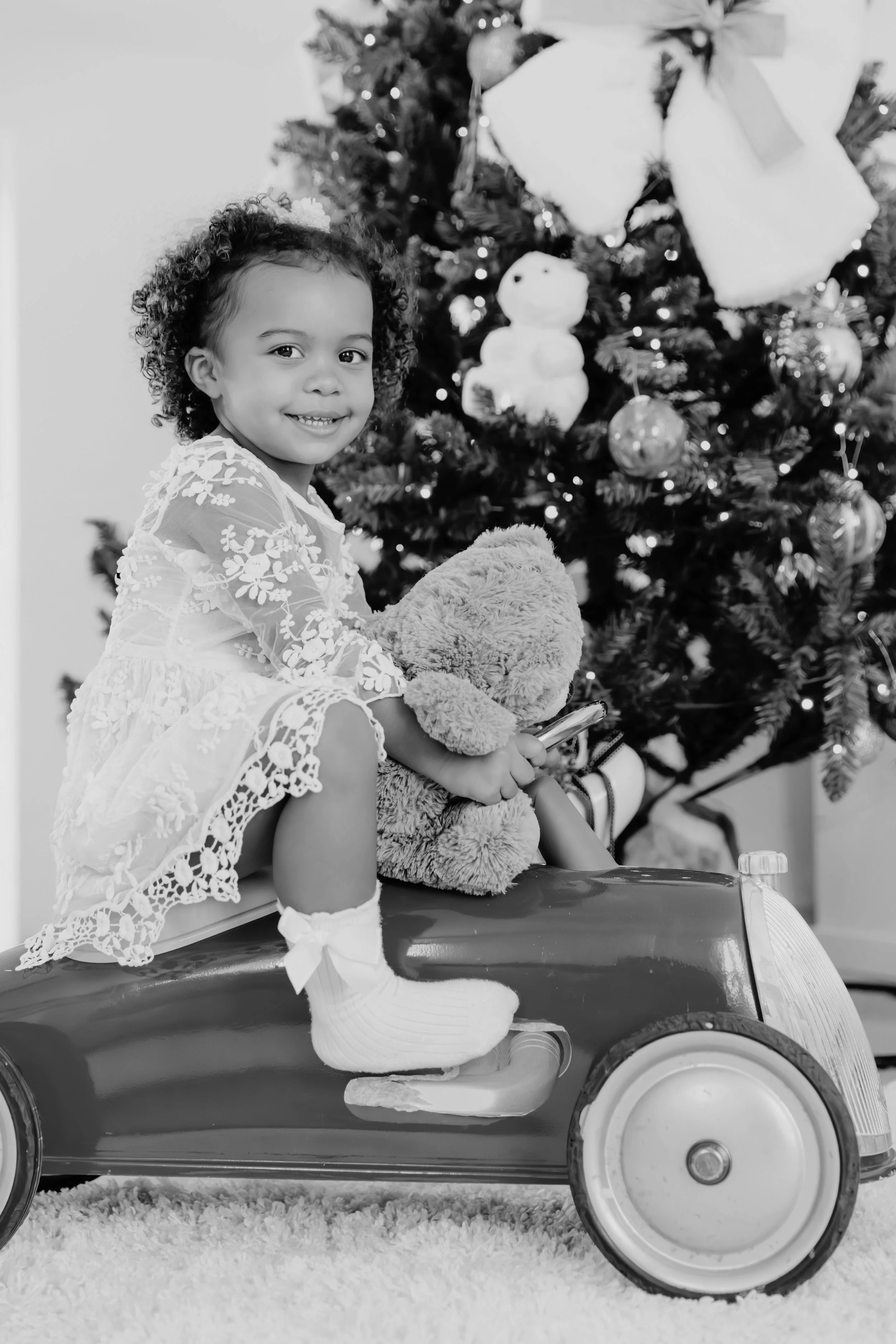 Une jeune fille souriante assise dans une voiture jouet avec un ours en peluche, devant un arbre de Noël décoré avec des boules, des chaussettes de Noël, et une boule de neige en peluche.