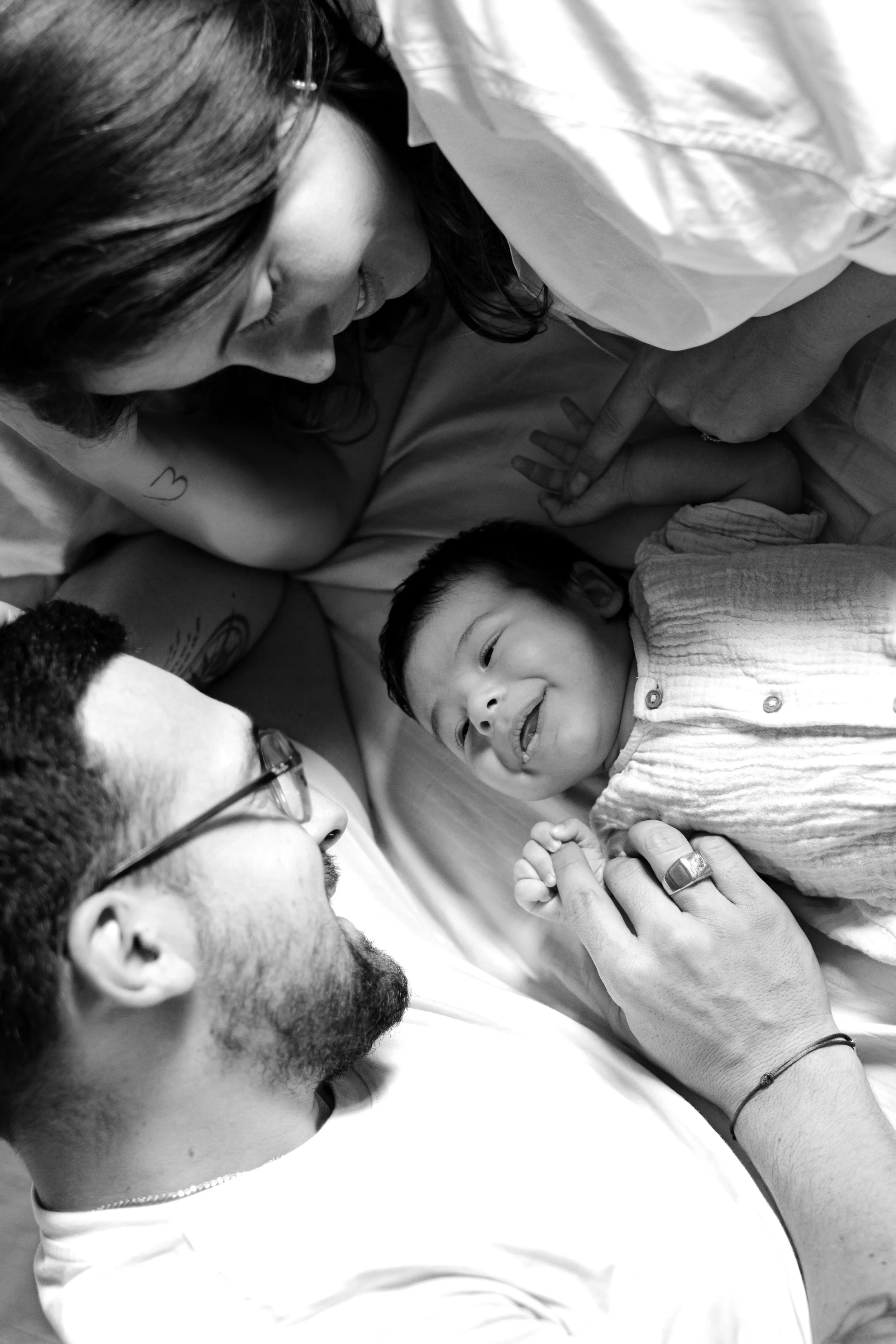 Une famille comprenant une femme, un homme avec des lunettes, et un jeune enfant, allongée dans un lit, partageant un moment affectueux.
