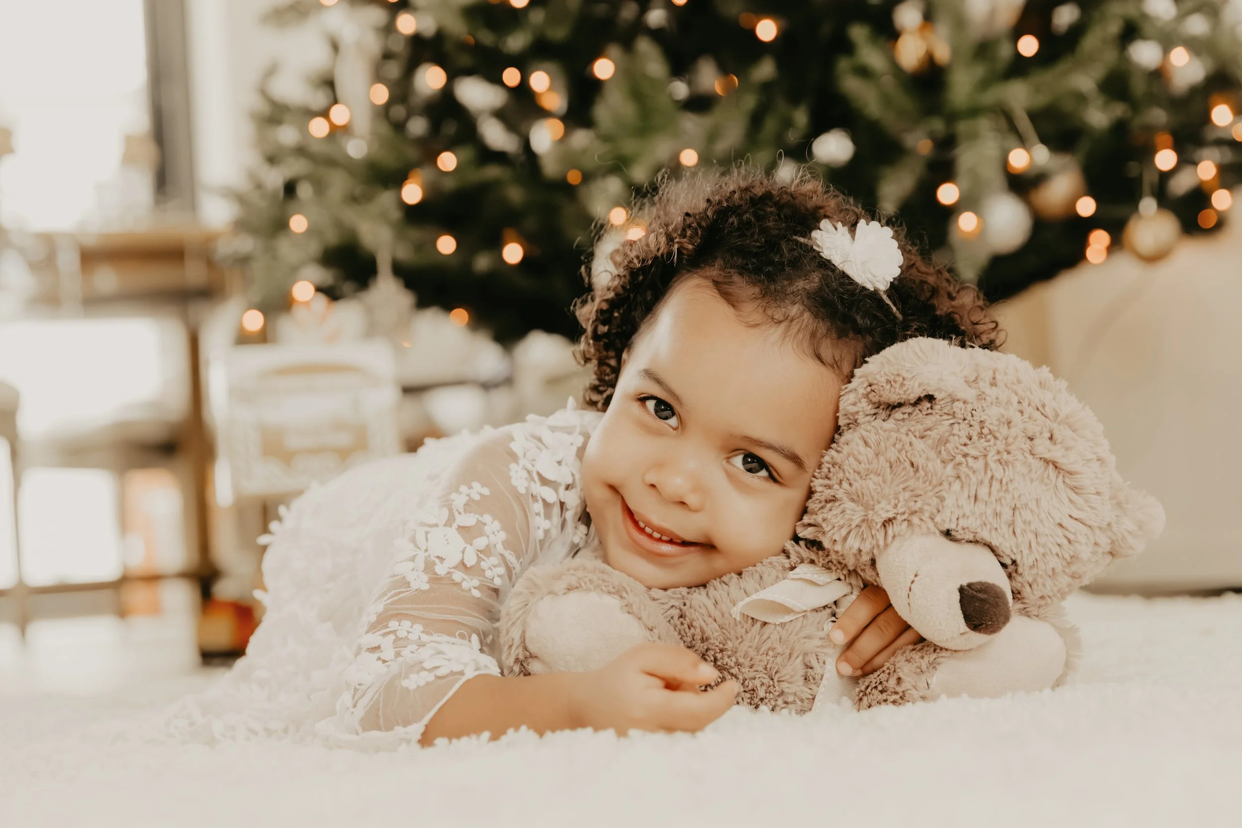 Une jeune fille souriante allongée sur un tapis blanc, tenant un ours en peluche, avec un sapin de Noël illuminé en arrière-plan.