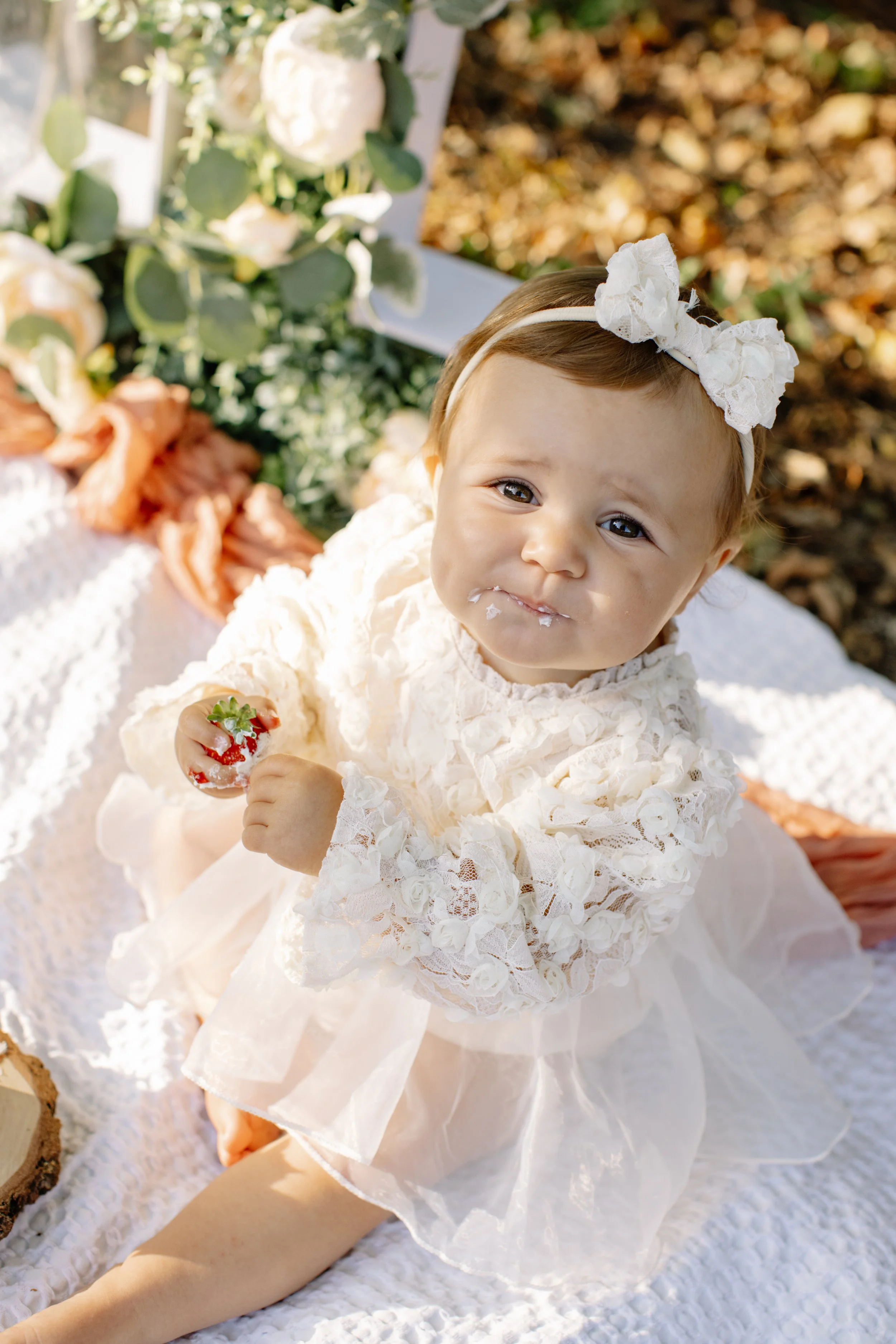 Une jeune fille assise sur une couverture en lin blanc, tenant une fraise, avec des décorations florales en arrière-plan, dans un cadre extérieur en automne.