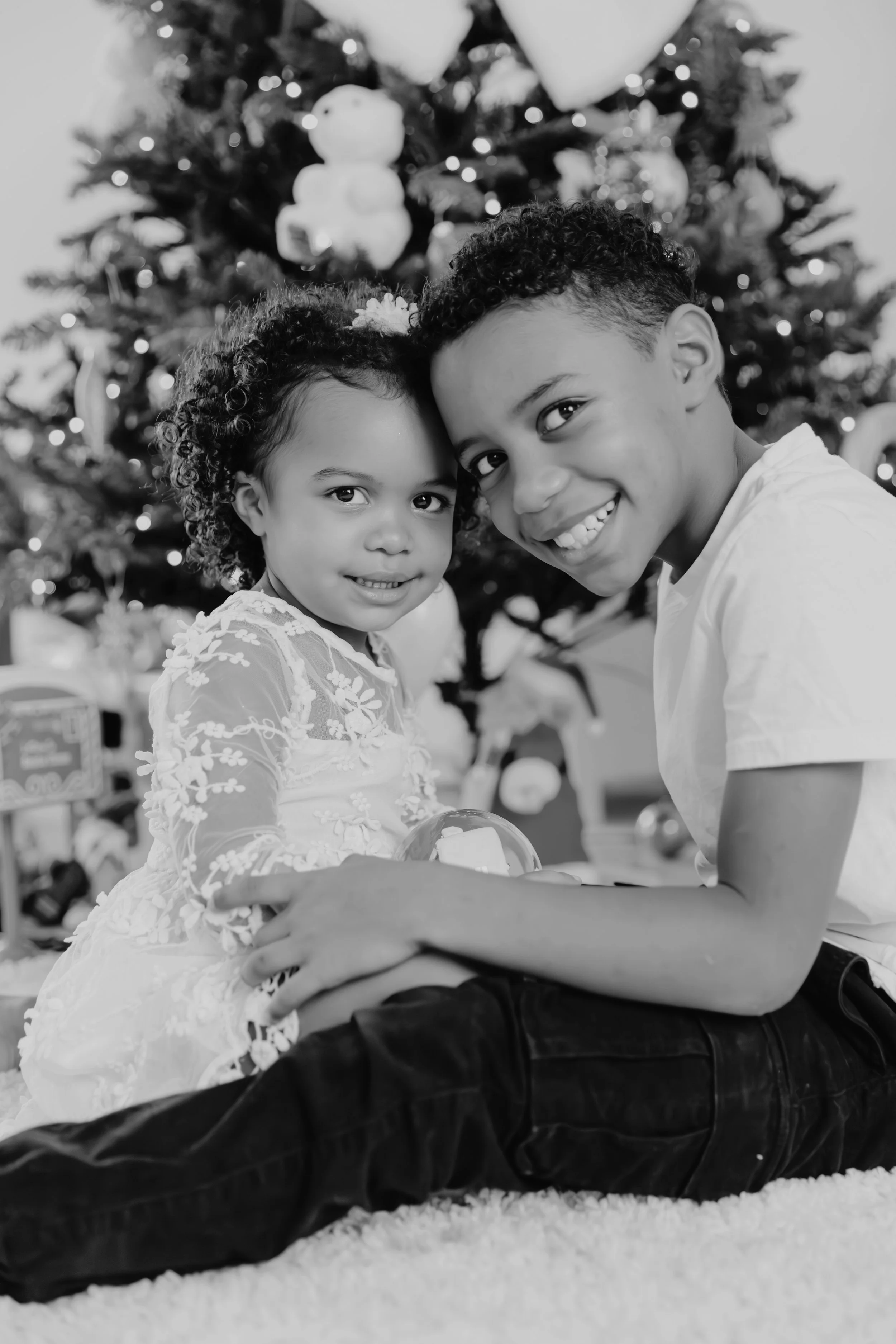 Deux enfants, une fille et un garçon, sourient devant un sapin de Noël décoré avec des guirlandes et des cadeaux, en noir et blanc.