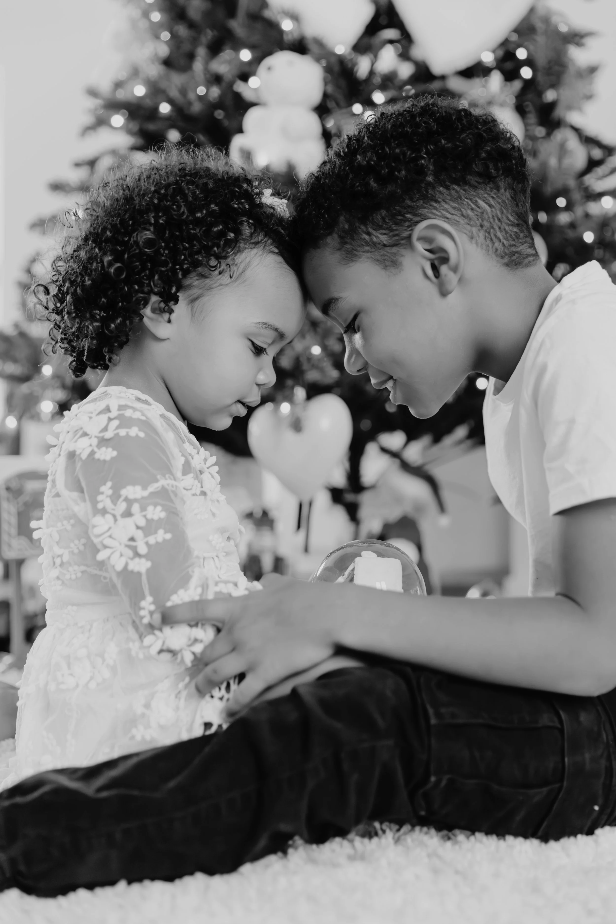 Deux enfants, un garçon et une fille, s'embrassent avec la tête inclinée l'un vers l'autre devant un arbre de Noël décoré.
