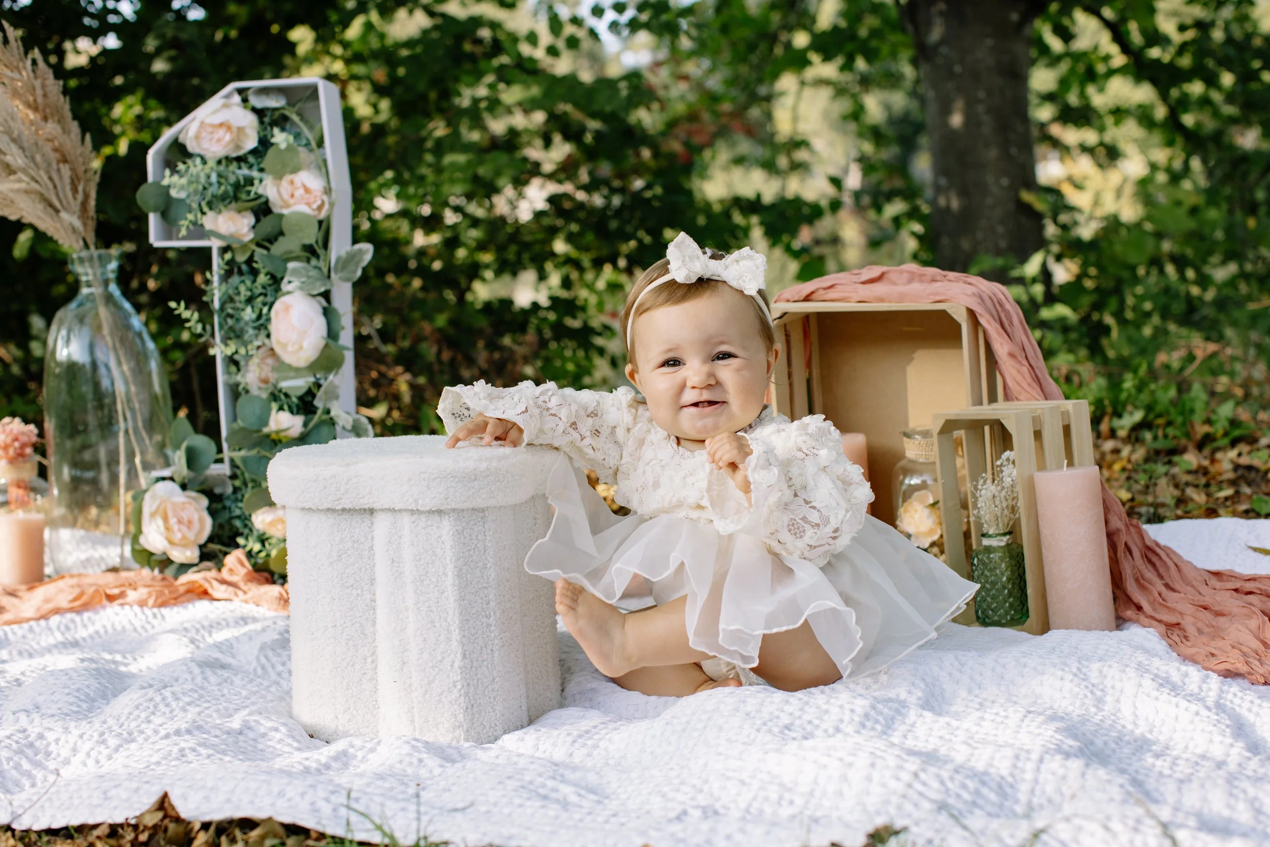 Jeune enfant en robe blanche, assise sur une couverture blanche, dans un décor extérieur avec fleurs et objets de décoration pour une photoshoot.