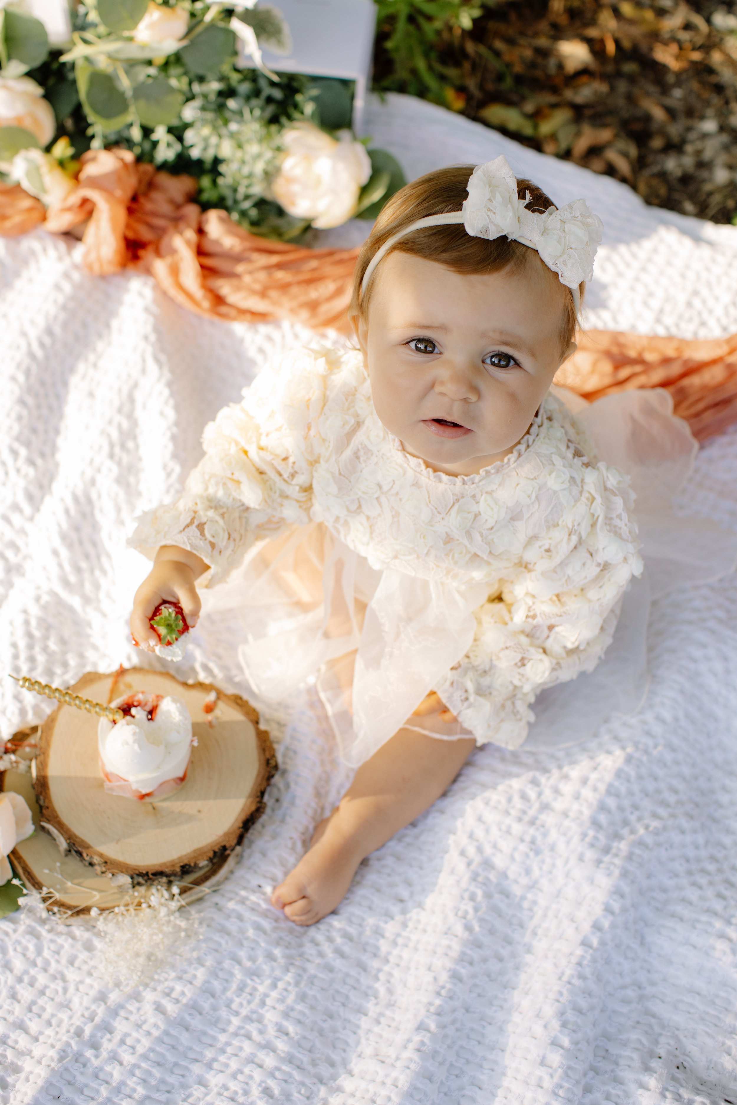 Bébé en robe blanche avec un bandeau blanc, jouant avec une fraise sur un gâteau en rondins en plein air sur une couverture blanche