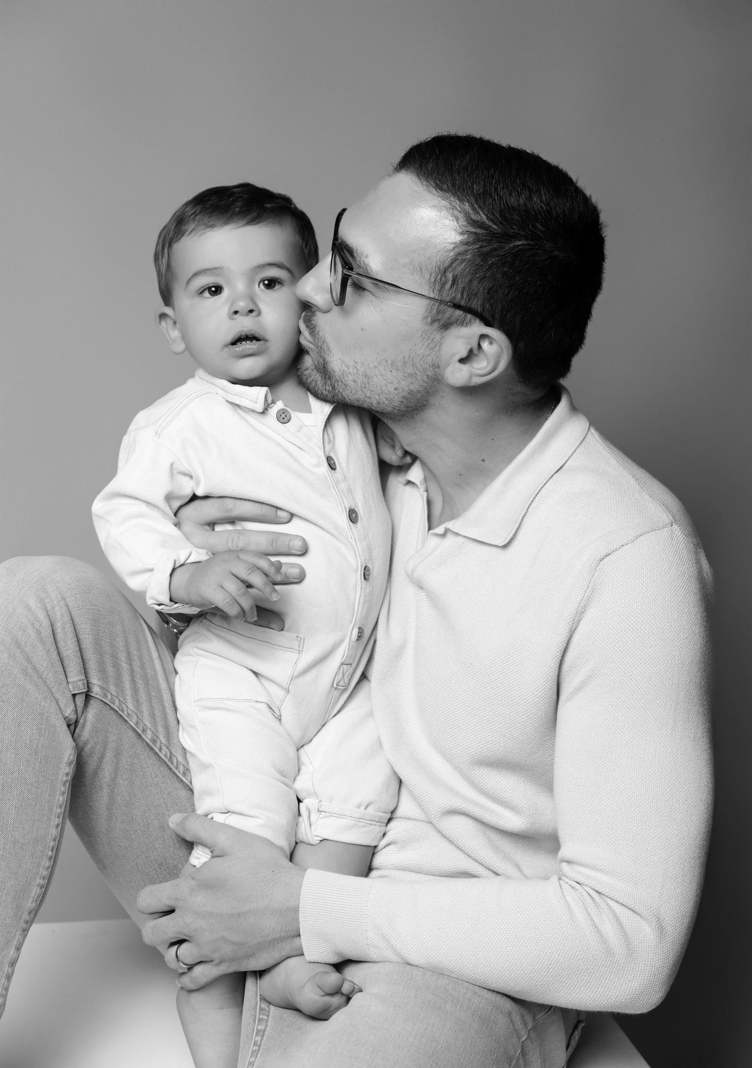 Un homme, porte des lunettes, embrasse tendrement un jeune garçon sur la joue. Le garçon regarde devant avec une expression neutre. La photo est en noir et blanc.