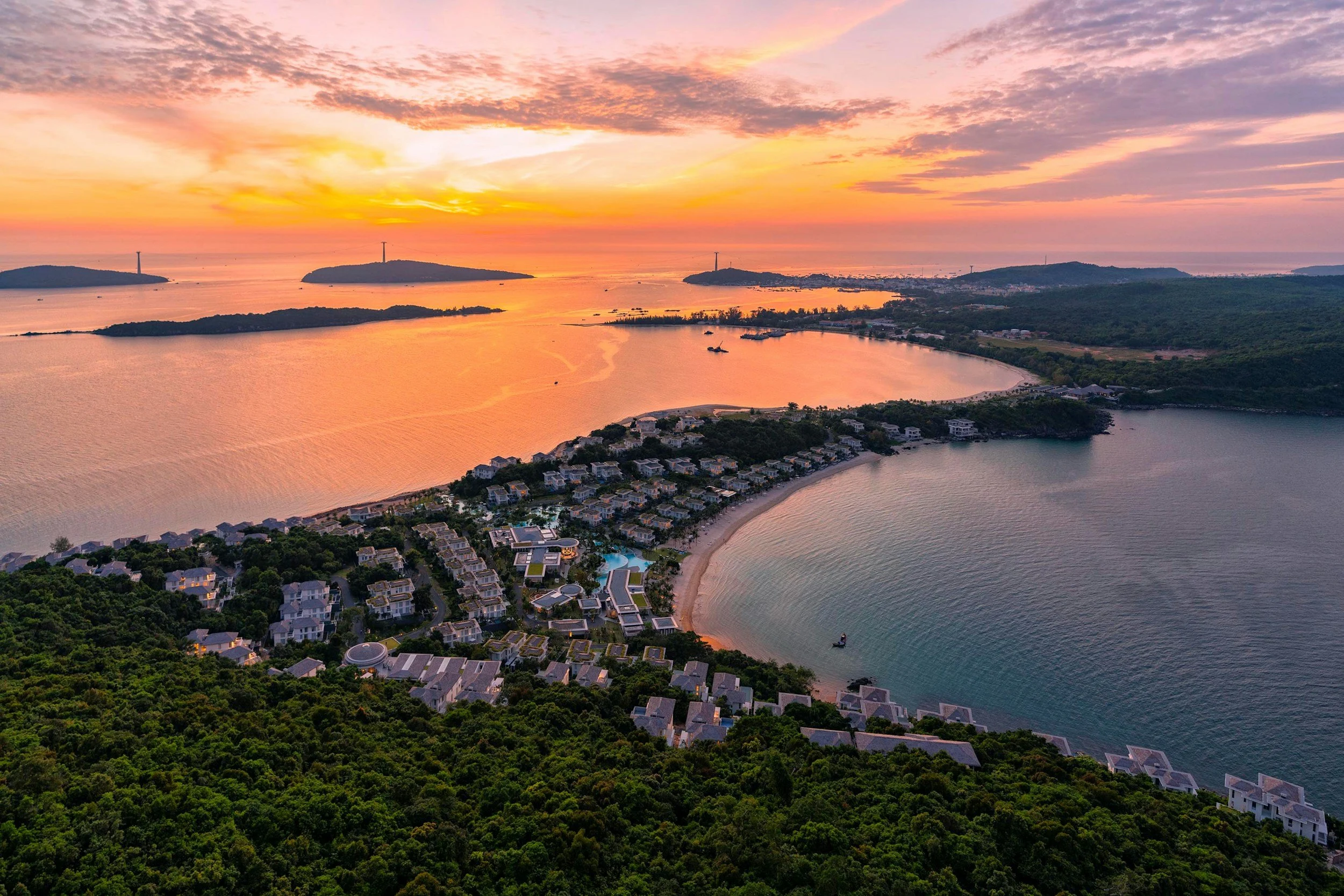 Aerial view of a coastal resort at sunset, showing a beach with houses, trees, and water with islands in the distance.