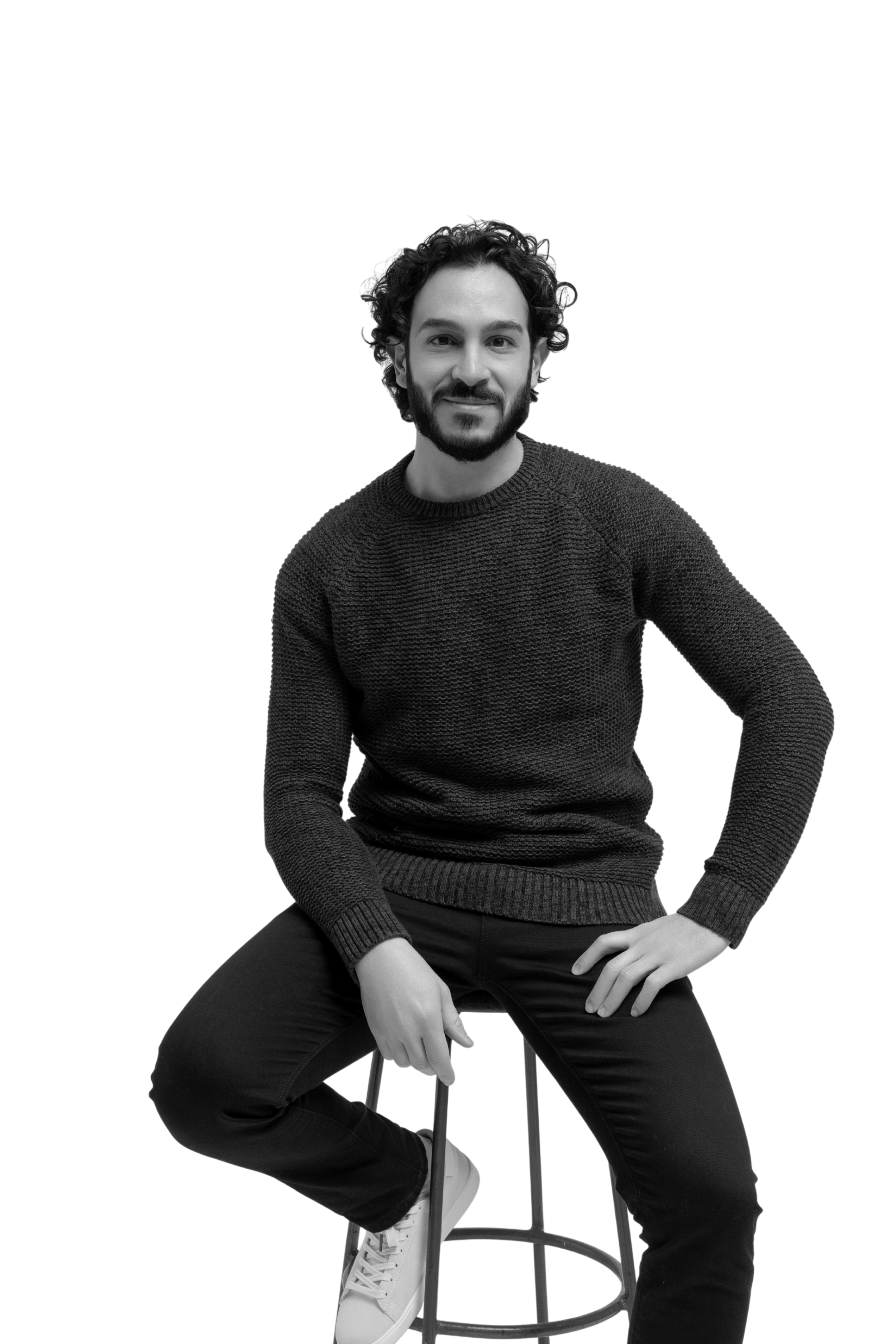 A young man with curly hair, beard, and light skin sitting on a stool, wearing a dark knitted sweater, black pants, and white sneakers, smiling at the camera.