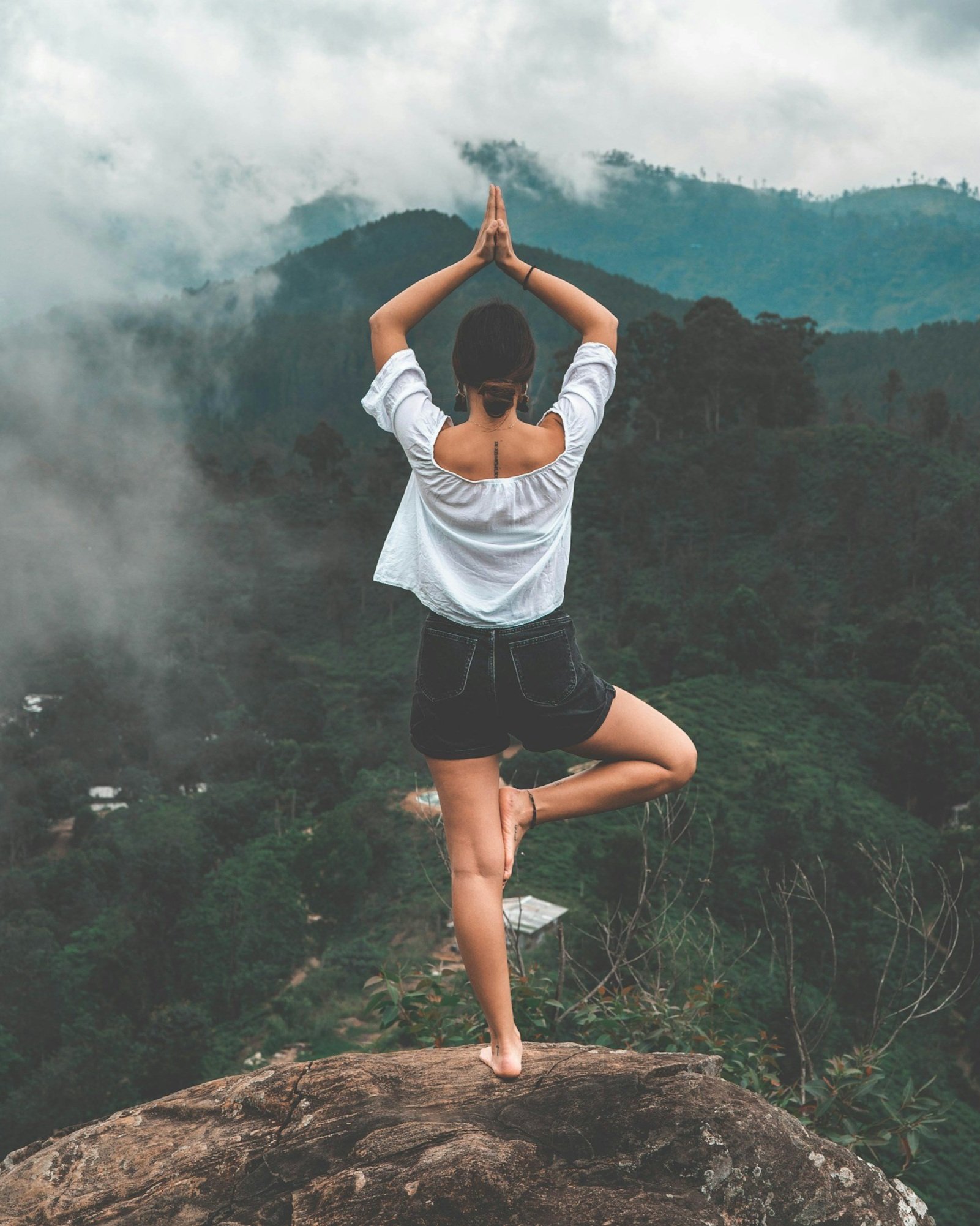 Woman practicing yoga on a rock ledge with a mountain landscape and cloudy sky in the background.