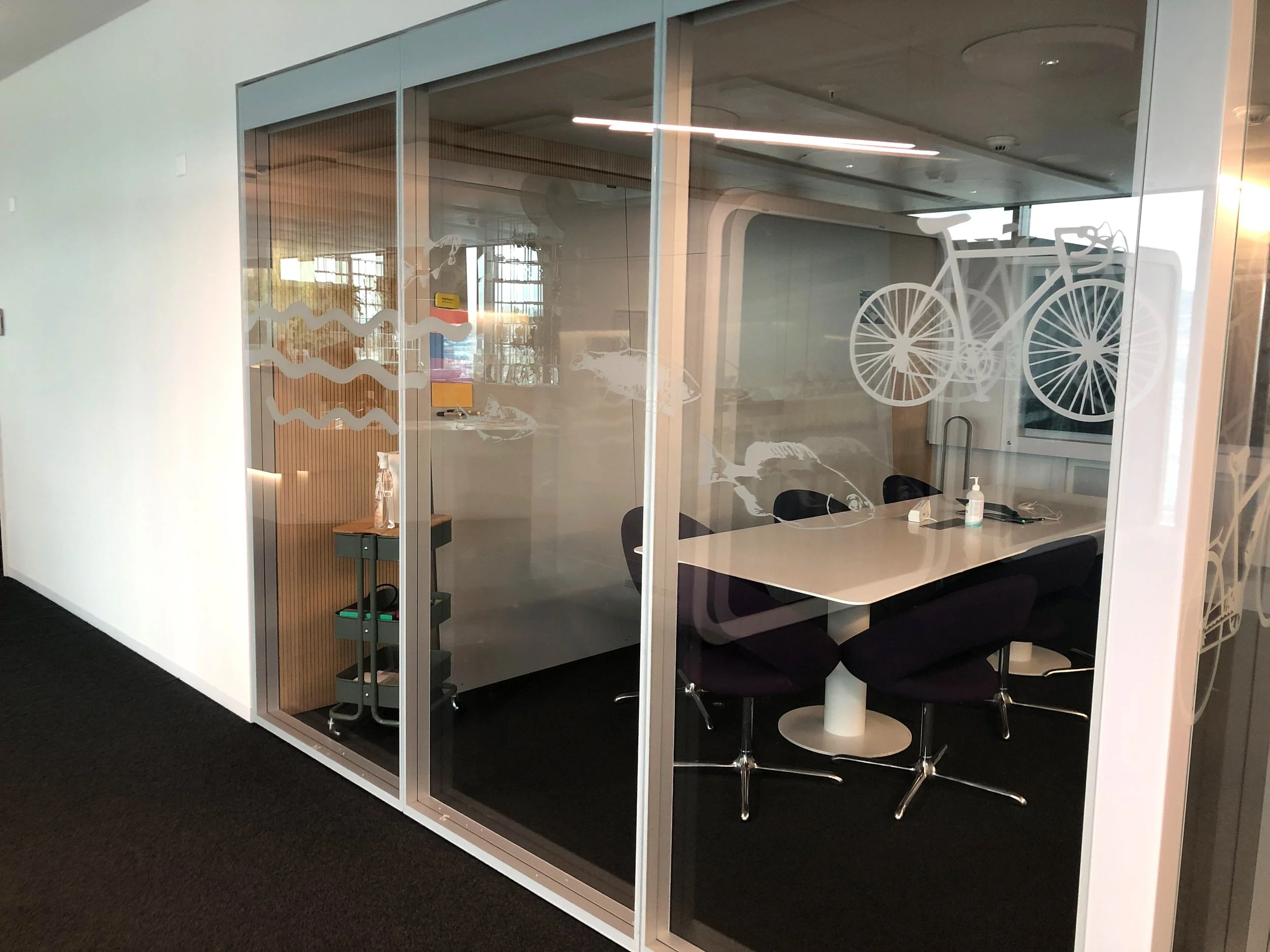 Moderner Besprechungsraum mit Glastüren, Fahrradaufkleber, einem Tisch, sechs Stühlen, einem Desinfektionsmittel und weiteren Büromaterialien.