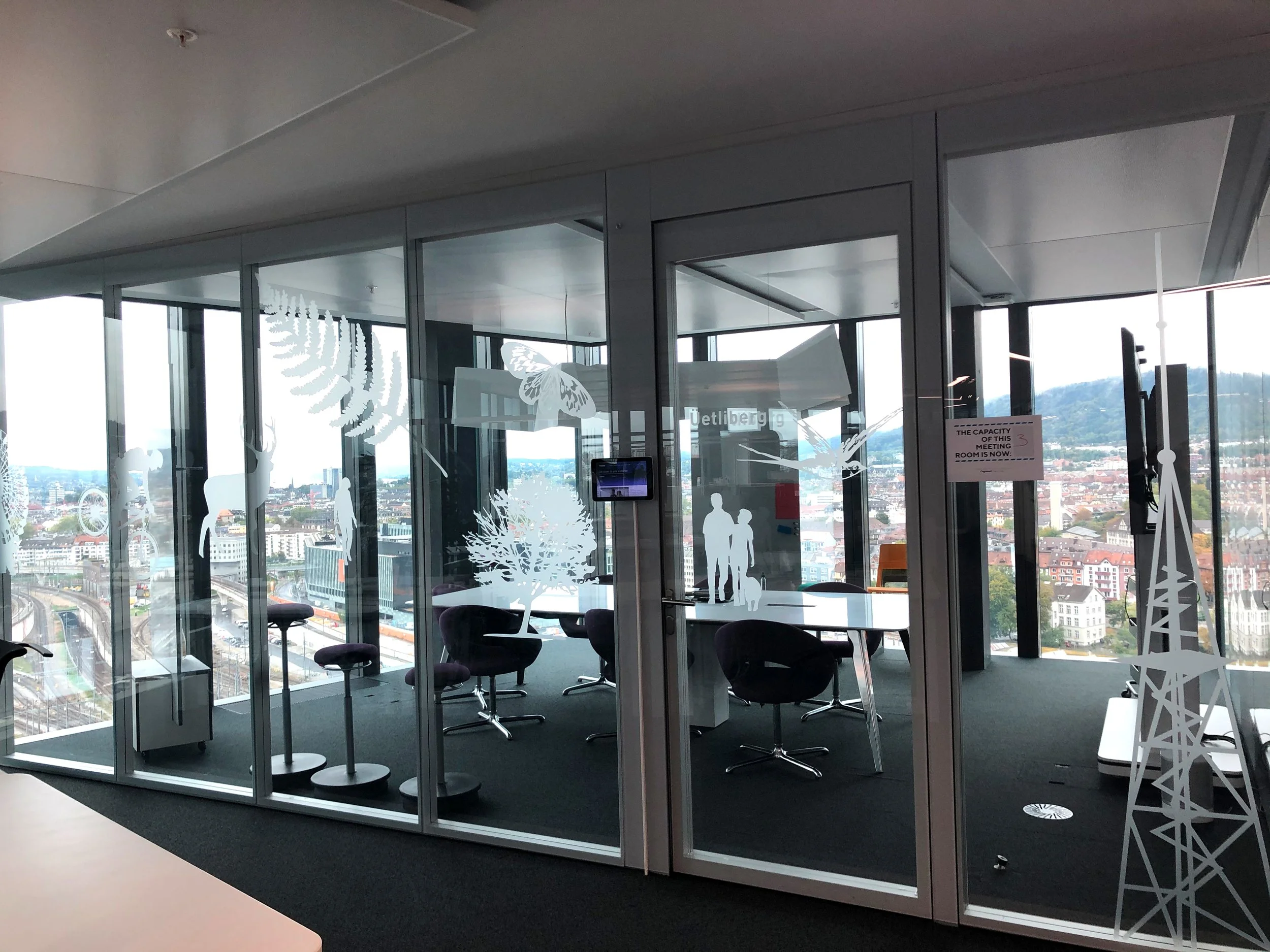 Innenraum eines modernen Büros mit großen Fensterflächen, die eine Stadt und Berge im Hintergrund sichtbar machen. Das Büro ist durch Glaswände abgetrennt und mit Dekorationen wie Baum- und Tierstickern versehen.