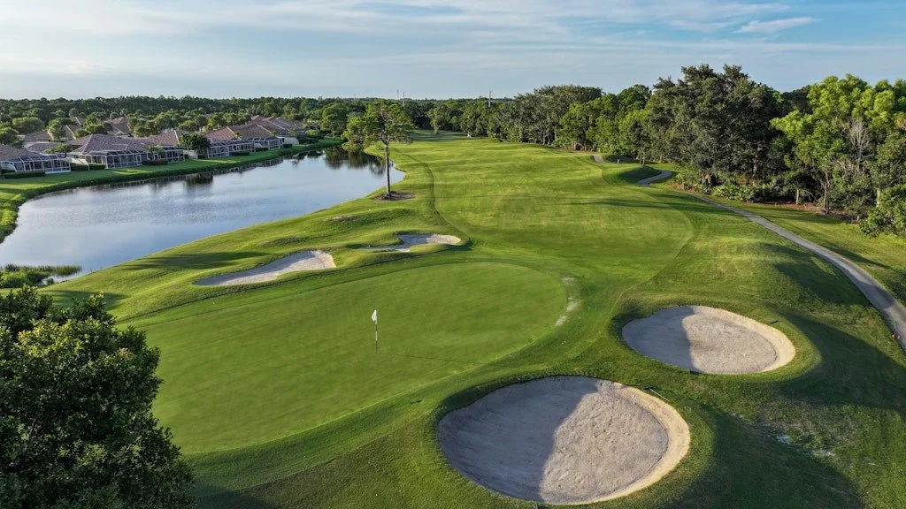 A golf course with a green and a flag, surrounded by sand bunkers, near a water hazard, with residential homes and trees in the background.