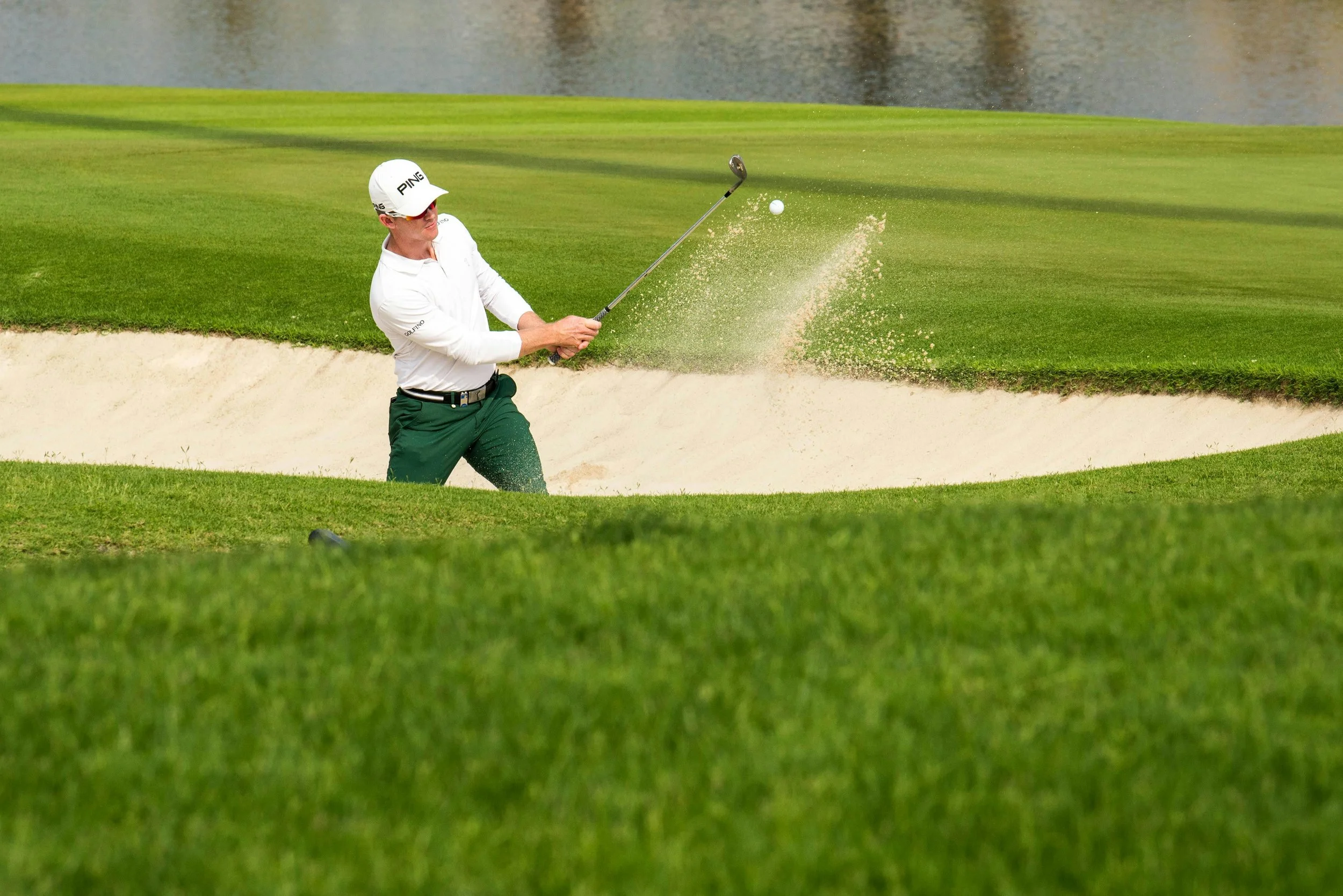 A golfer in a white shirt, green pants, and a white cap is hitting a golf ball out of a sand trap bunker on the golf course, with water visible in the background.