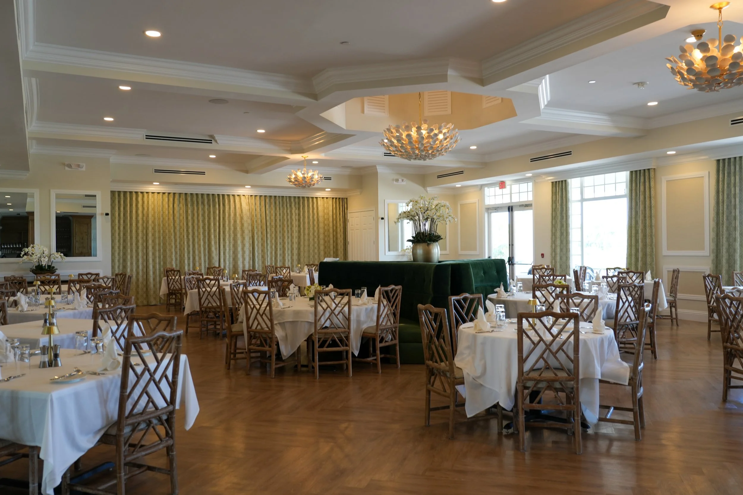 Elegant restaurant dining area with round tables covered in white tablecloths, set with glassware and napkins, wooden chairs with crisscross backs, large floral centerpieces, and yellow patterned curtains. The room has a high ceiling with crown molding, modern chandeliers, and large windows letting in natural light.