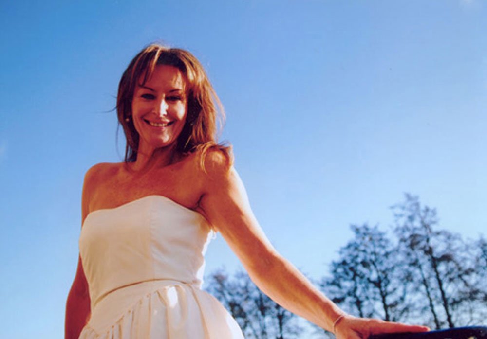 Woman smiling outdoors during the daytime with clear blue sky and trees in the background.