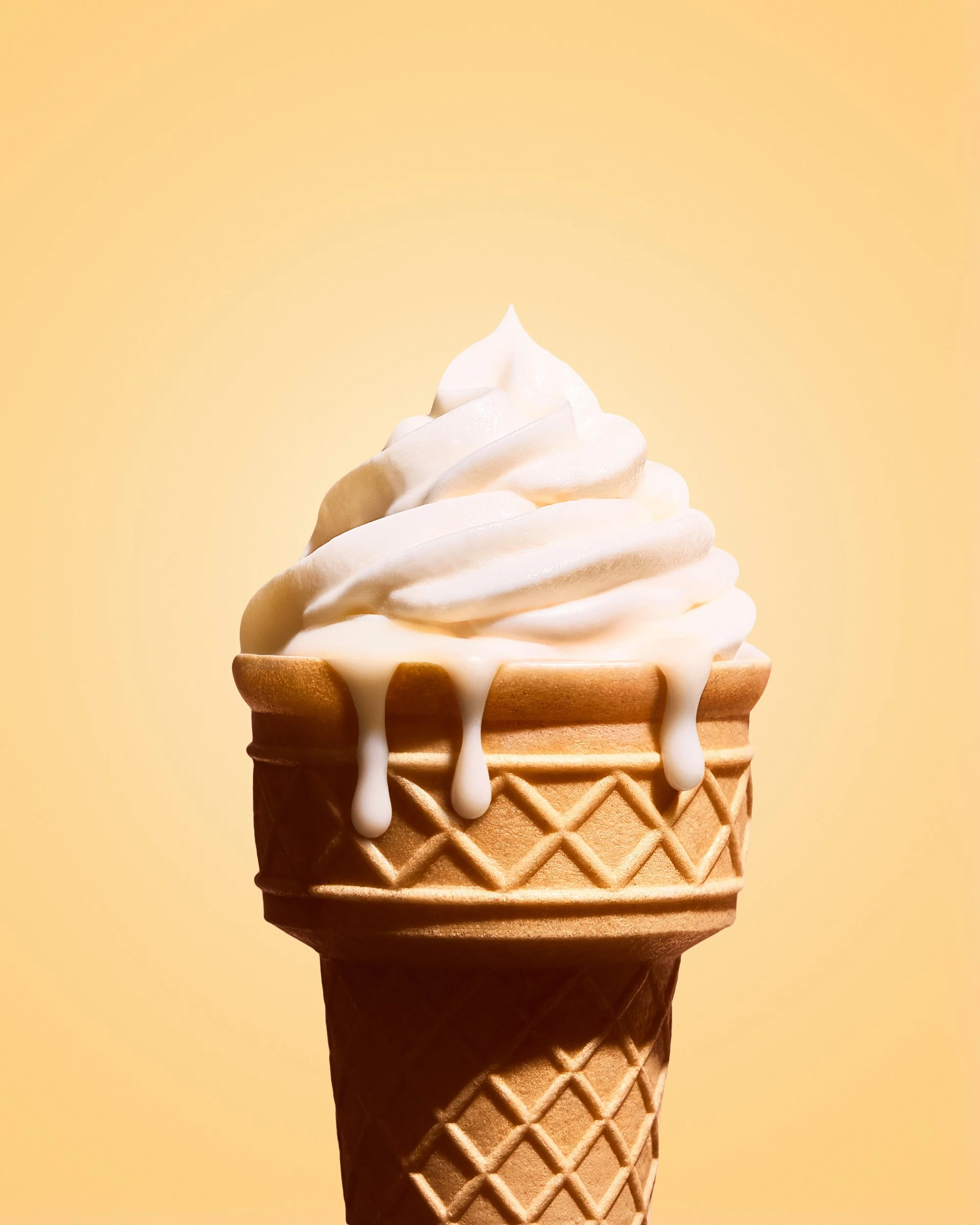 Close-up of a vanilla ice cream cone with a waffle pattern, with white melted ice cream dripping down the sides, against a plain peach-colored background.