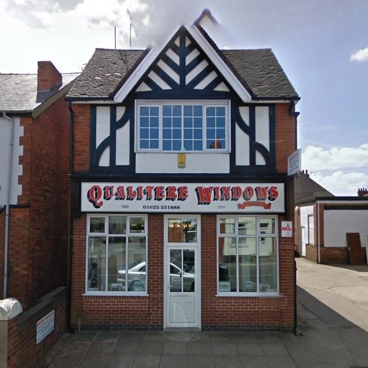 A two-story brick building with a sign that reads 'Qualtere Windows' and a phone number, featuring large front windows and a white door, serving as a window shop or showroom