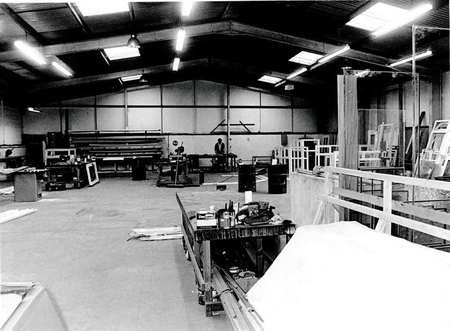 Black and white photo of a woodworking or carpentry workshop with various tools, materials, and wood frames arranged around the space, including a person standing in the middle.