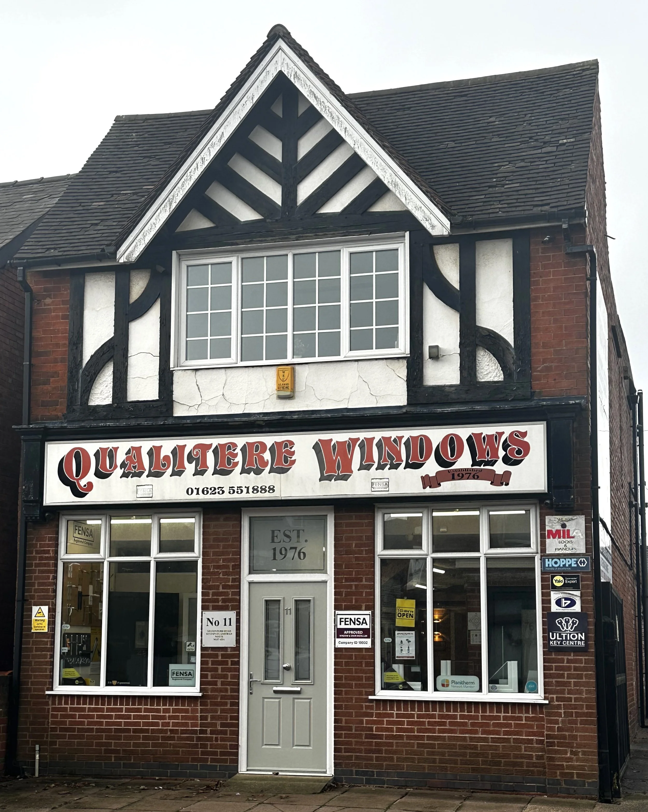 A two-story brick building with a black and white Tudor-style upper facade housing "Quali Tere Windows" store, established in 1976, with large glass windows on the ground floor displaying various sign stickers, and a gray door with a sign that says "No 11" and "EST 1976" on the upper window. There are multiple logos of window brands on the right side and security system signs near the door.