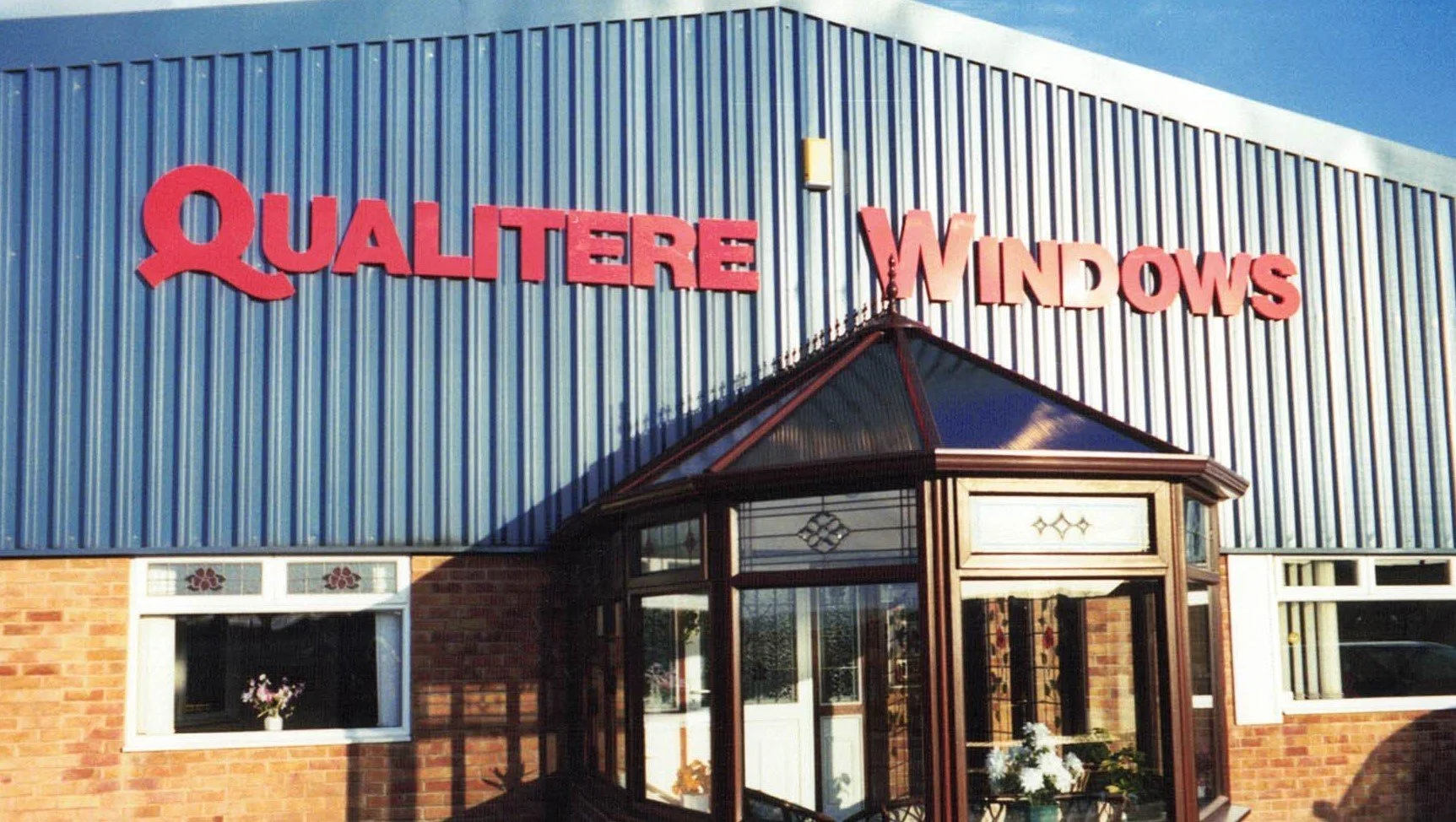 The building has a blue metal exterior with red letters that read 'QUILTERE WINDOWS.' There is a glass entrance with a small porch and windows, some decorated with flowers.