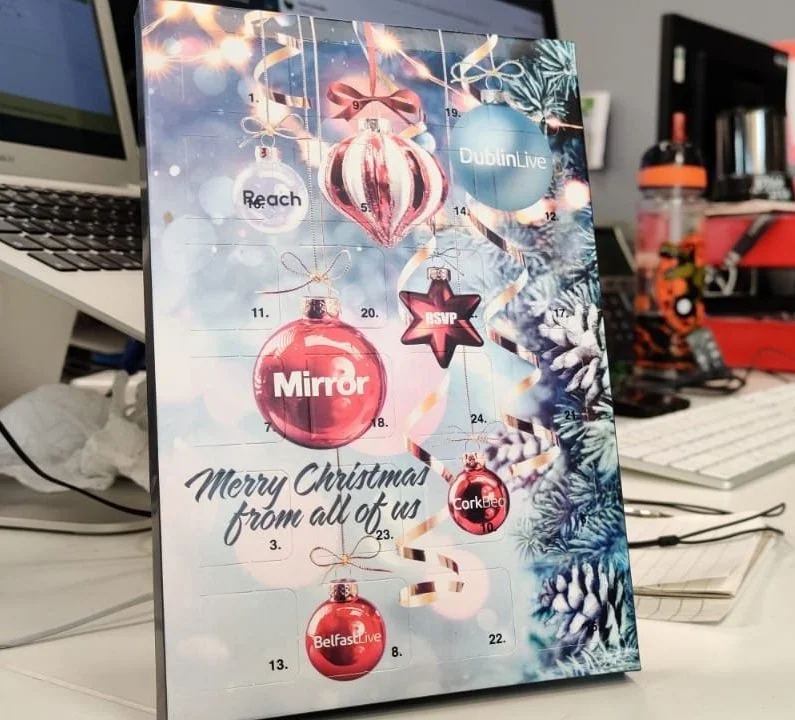 An Advent calendar with Christmas ornaments and lights, displaying holiday greetings from all of us, with numbered doors for each day.