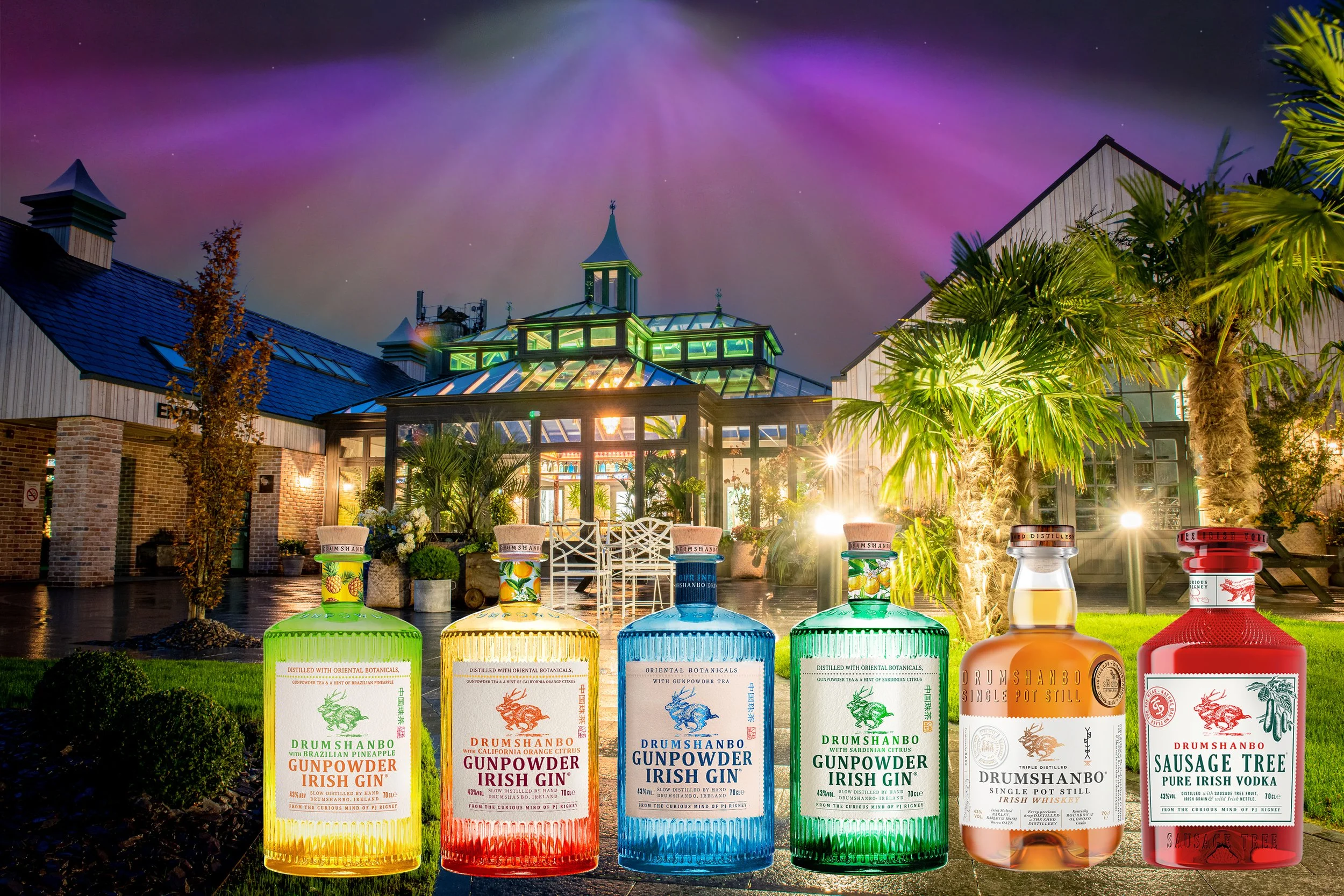 A row of six bottles of Irish spirits in front of a lit greenhouse and garden at night, with a colorful sky overhead.