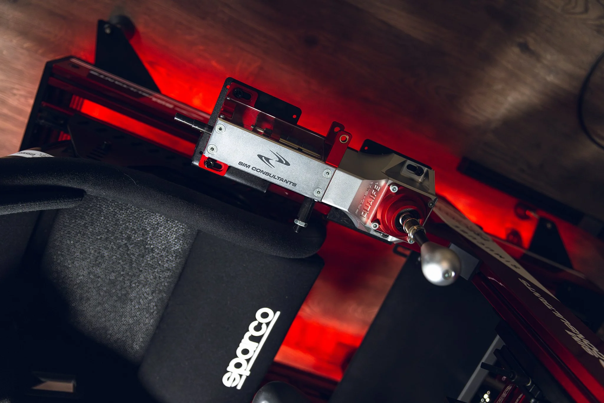 A racing cockpit simulator setup with a Sparco racing seat, a red and silver Simu Consultants motion platform, and a Porsche branded component, all illuminated by red lighting.