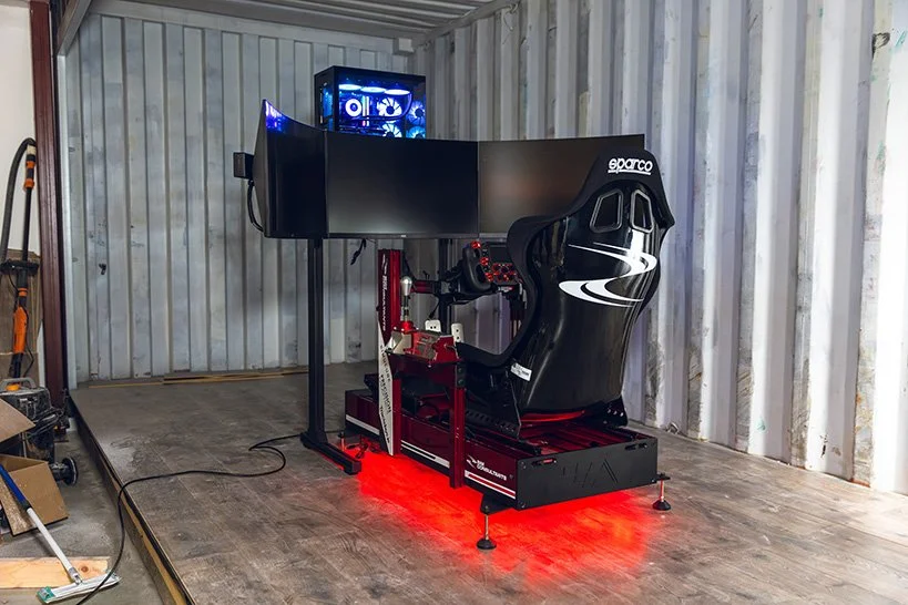 A racing simulator setup featuring a black Sparco racing seat, a racing wheel and pedal set, dual monitors, and a gaming PC with blue LED lighting, all on a wooden platform in a garage or storage space.