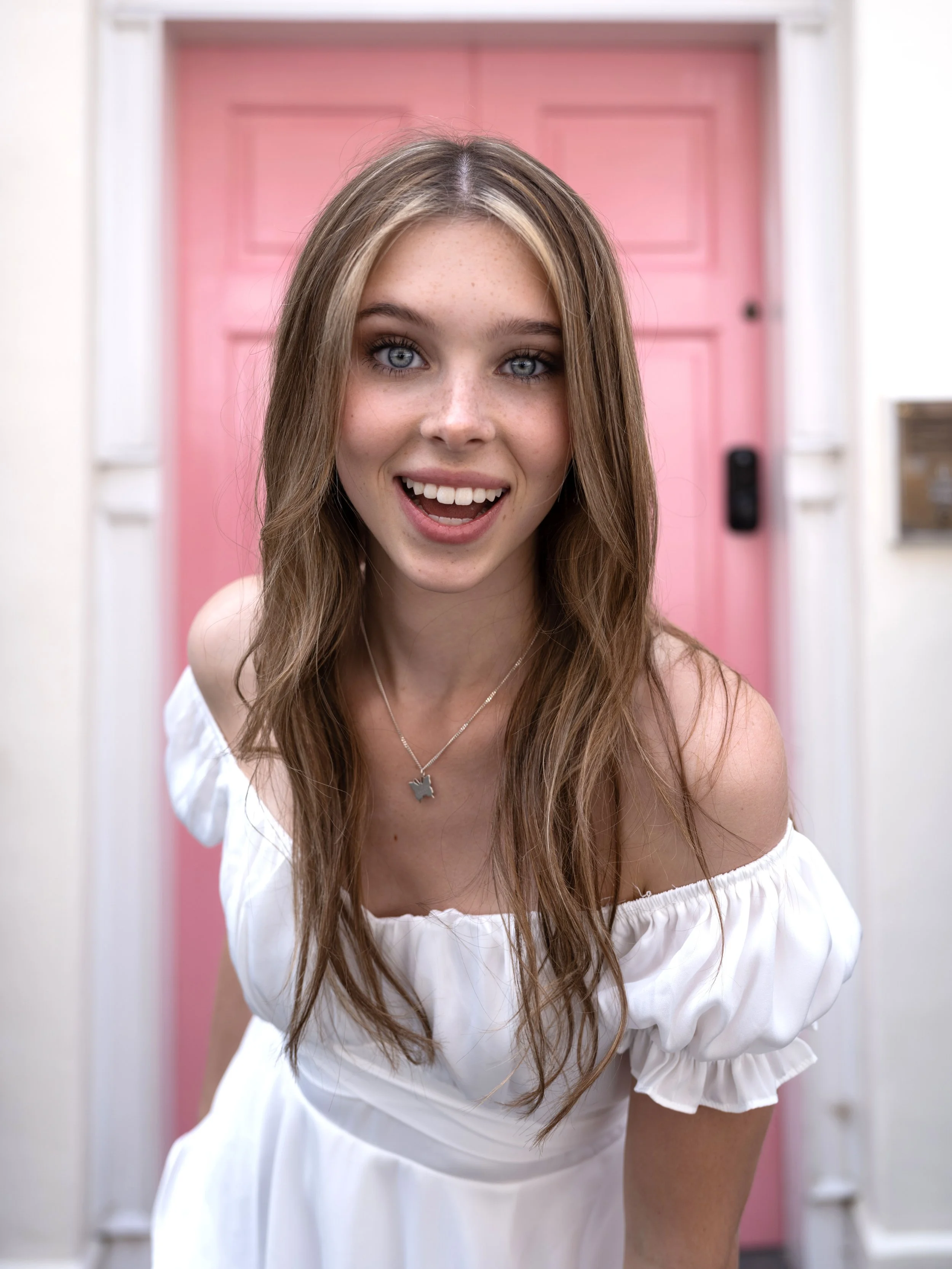 A portait of a girl smiling and leaning forwards wearing a white dress with a pink door in the background.