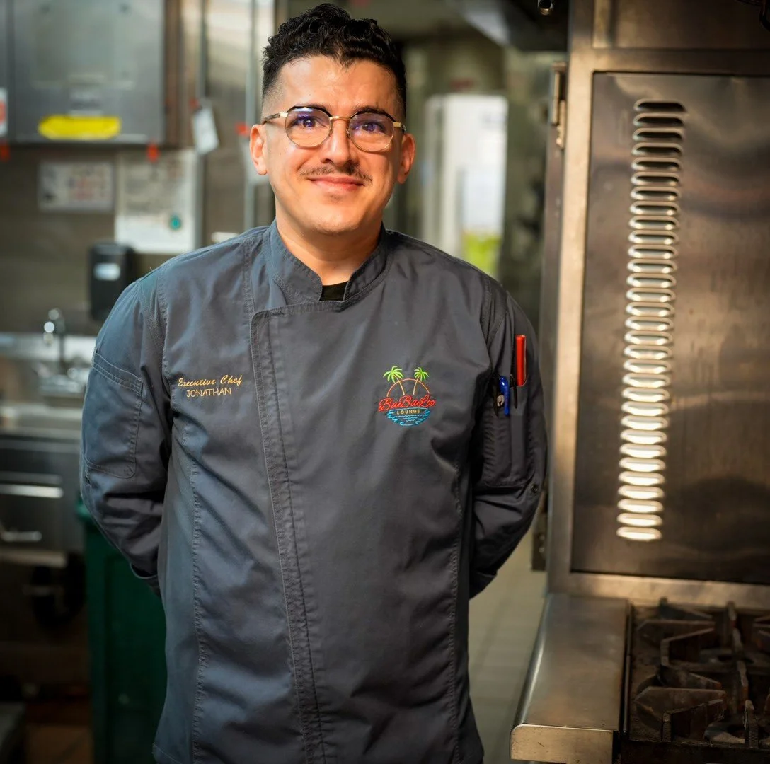 Portrait of a young male chef wearing a white chef's coat, a tall white chef's hat, and a medal around his neck, smiling with arms crossed against a dark background.