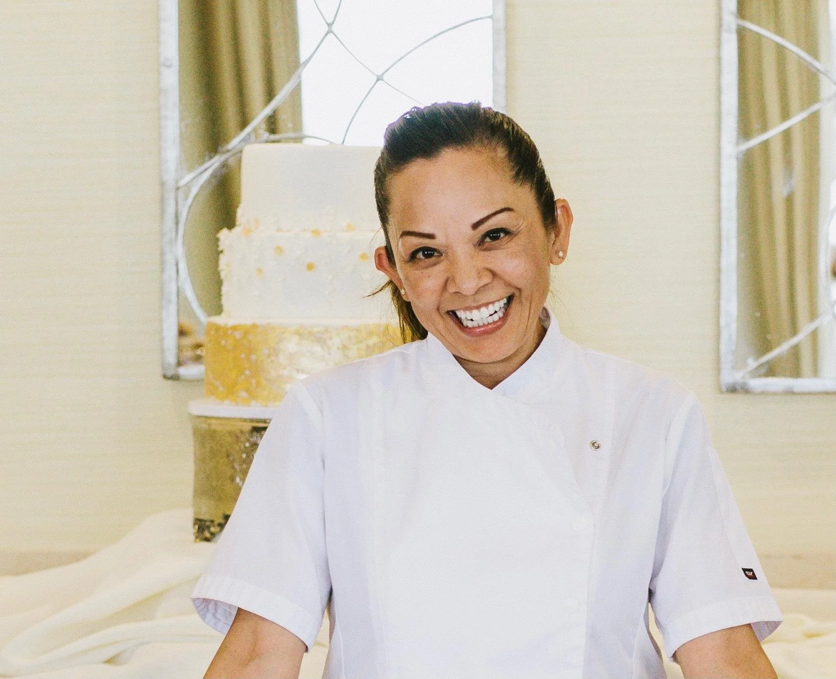 A woman in a white chef's coat smiling in front of a three-tier wedding cake with white and gold decoration, with a mirror and curtain background.