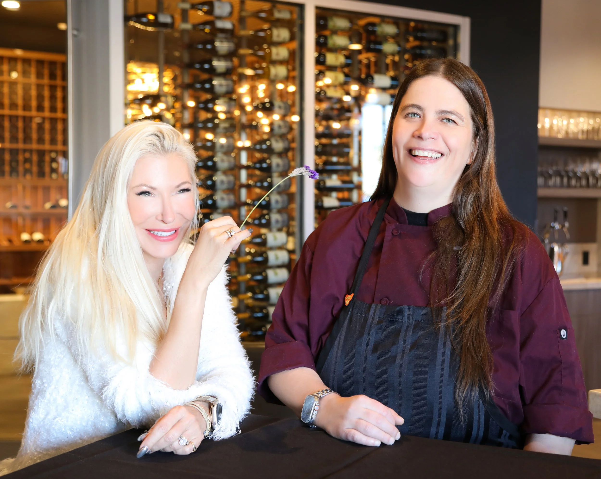 A woman with long blonde hair and a white sweater sitting at a table, smiling and holding a purple flower near her face, next to a woman with long brown hair wearing a burgundy chef's jacket and a black apron, smiling in a restaurant or wine cellar setting with a wine rack in the background.
