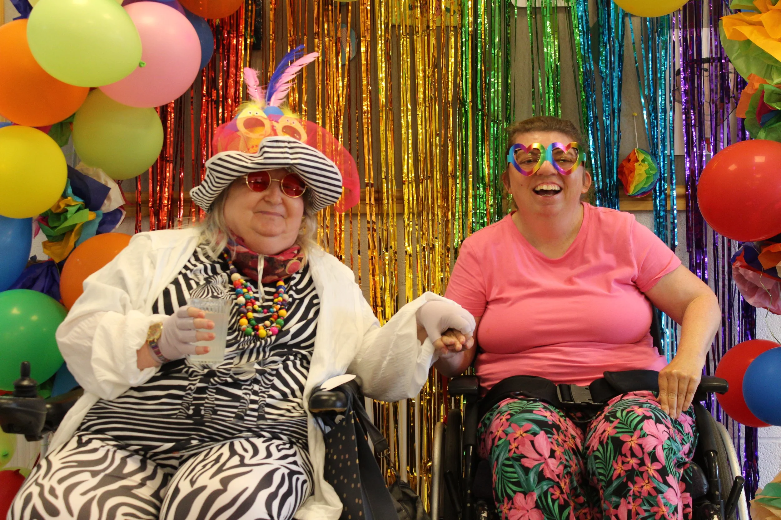 Two women sat in wheelchairs wearing bright clothing and sunglasses and smiling