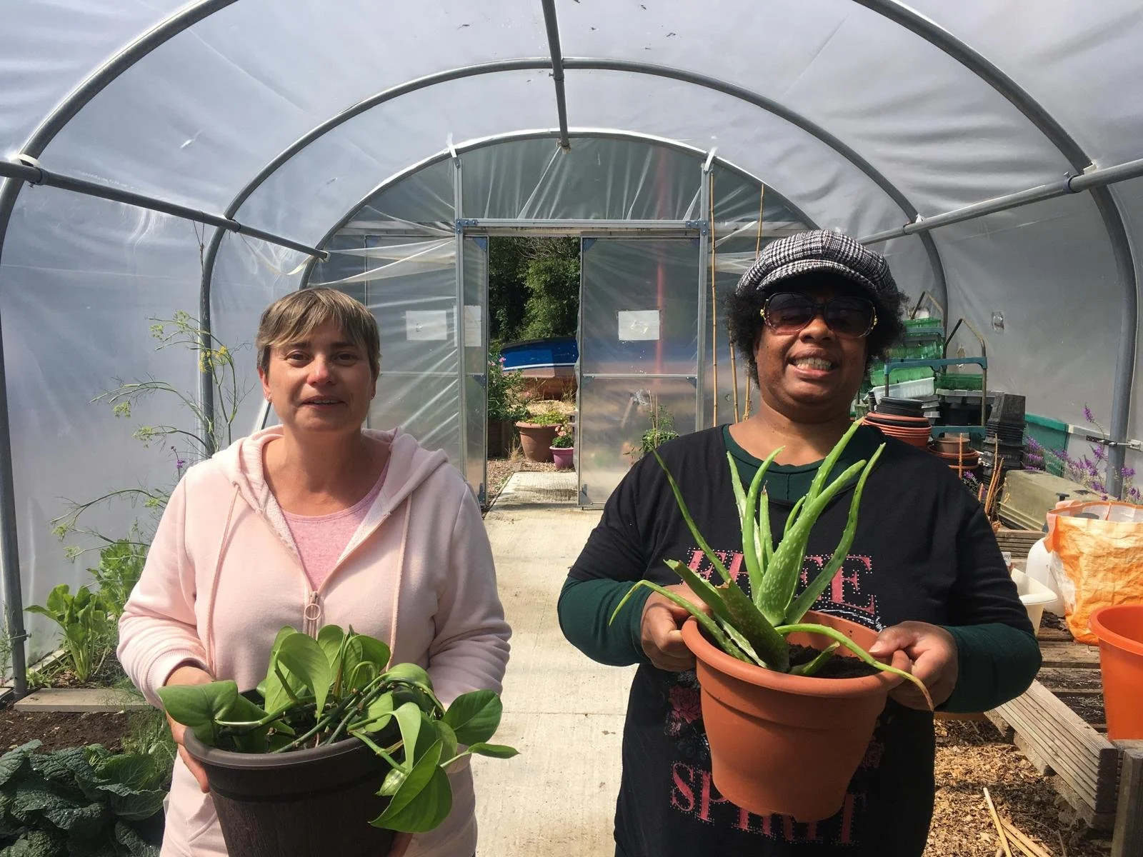 Two women holding plants in pots standing in a polytunnel doing gardening activities