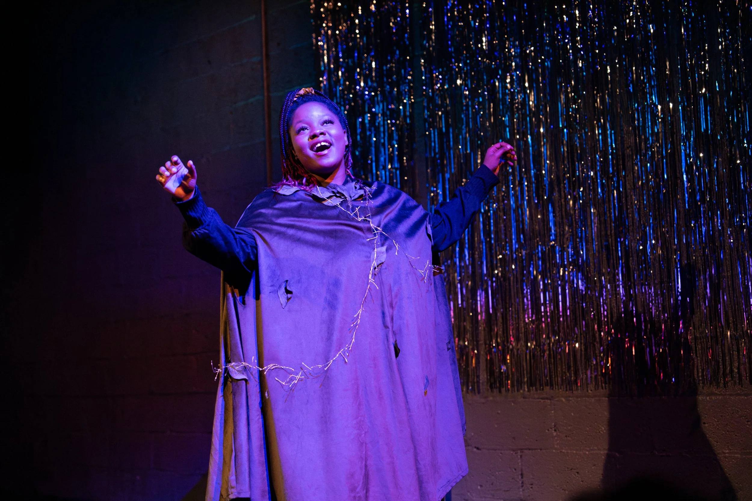 A young girl performing on stage with her arms outstretched wearing a purple robe and a string of lights, illuminated by colorful stage lighting, with a shiny curtain backdrop.