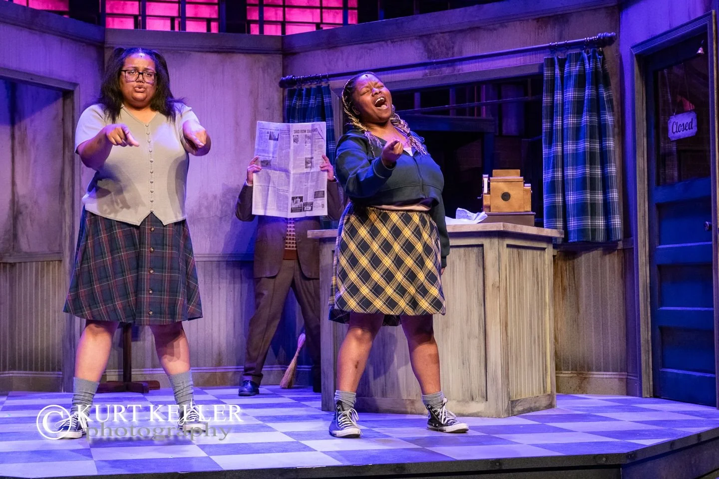 Theater stage scene with three women, one in a plaid skirt and gray top, singing passionately; another in a plaid skirt and brown blazer reading newspaper with a broom behind her; and a third woman in a gray cardigan, plaid skirt, and glasses making 
