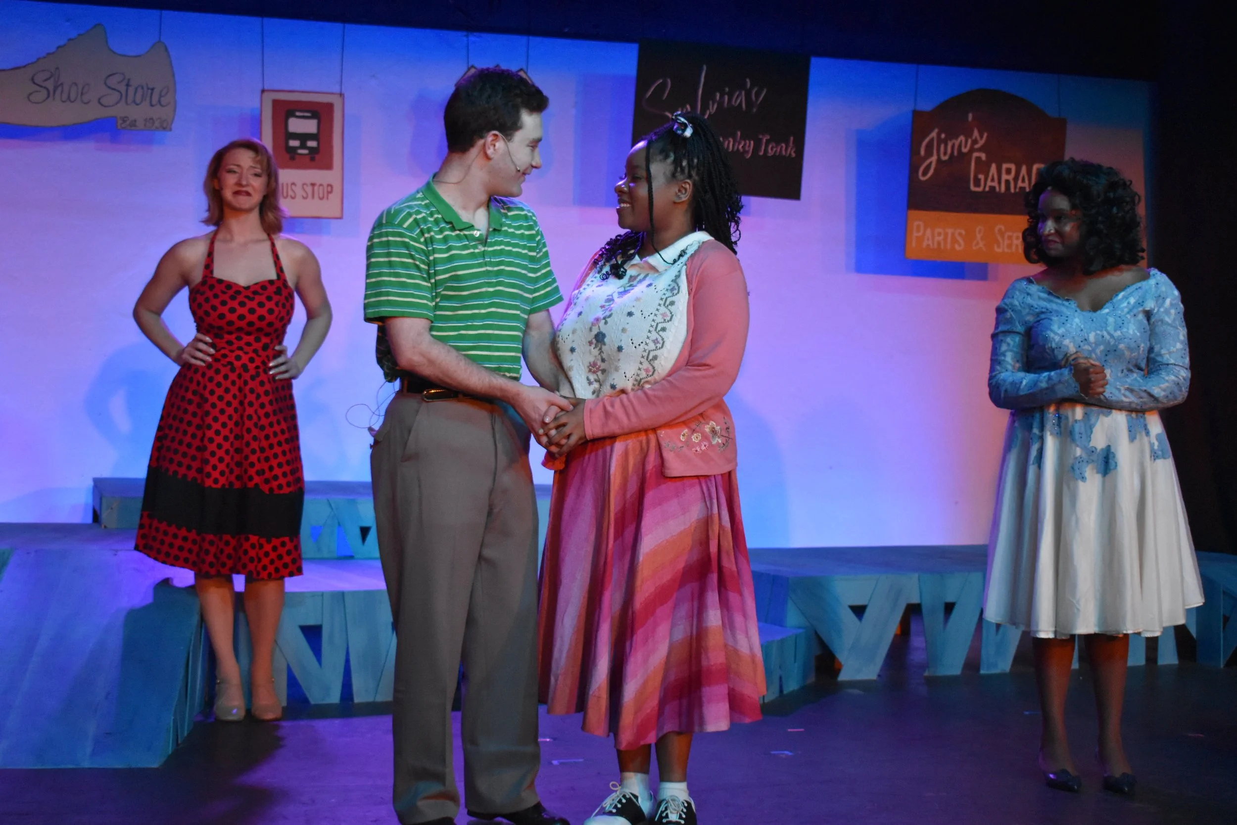 The scene is from a stage play with four women. Two women are in the center, holding hands and looking at each other, engaged in a moment. The woman on the left wears a green striped shirt and brown pants; the woman on the right wears a pink cardigan