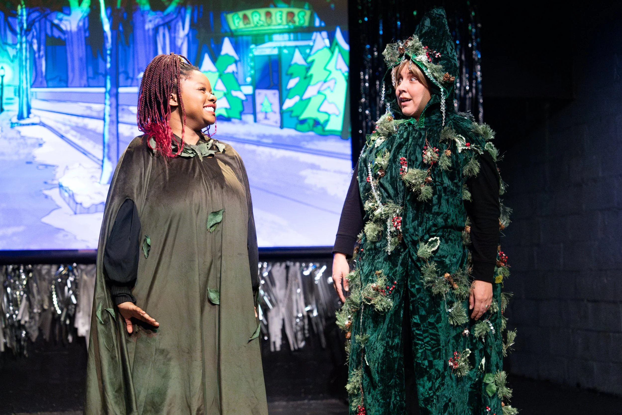 Two women dressed as Christmas trees, standing in front of a colorful animated backdrop of a forest with trees and a sign that says "Farbers". They are engaged in a conversation, smiling and looking at each other.