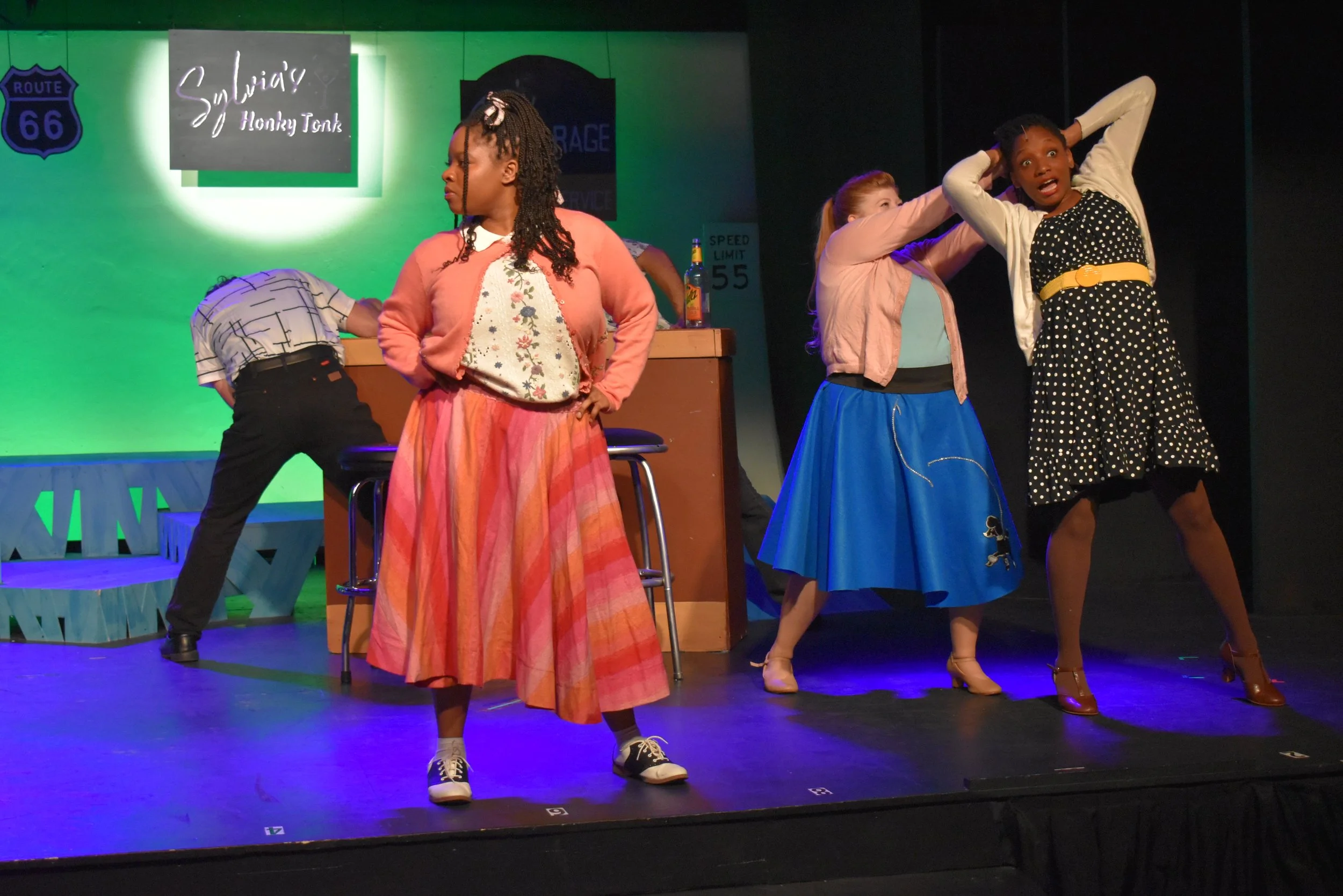 Four women in colorful vintage clothing on a stage, acting in a theatrical play with a bar set in the background, including signs for Route 66 and a speed limit. One woman stands with hands on her hips looking away, another adjusts her hair, and two 