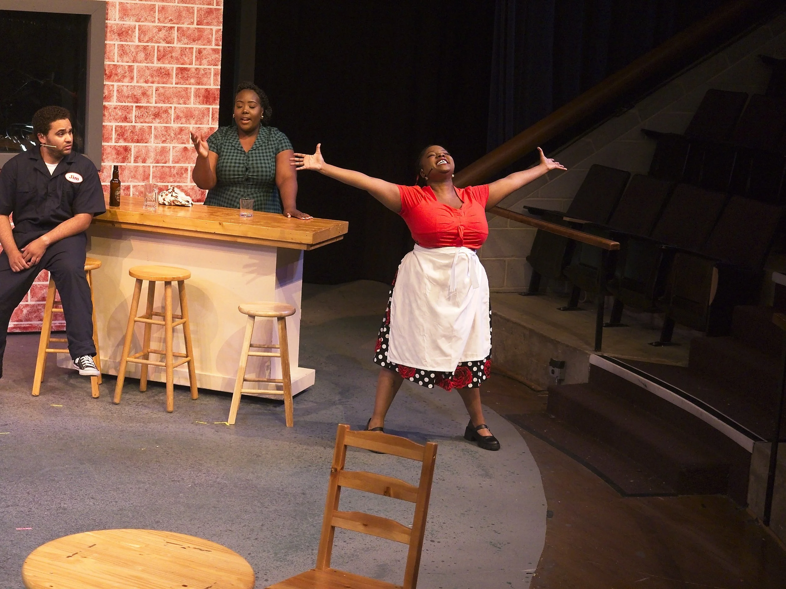 Three actors performing on stage, one woman singing with arms outstretched, two other actors listening, set to resemble a bar or restaurant with a countertop, stools, and brick wall backdrop.
