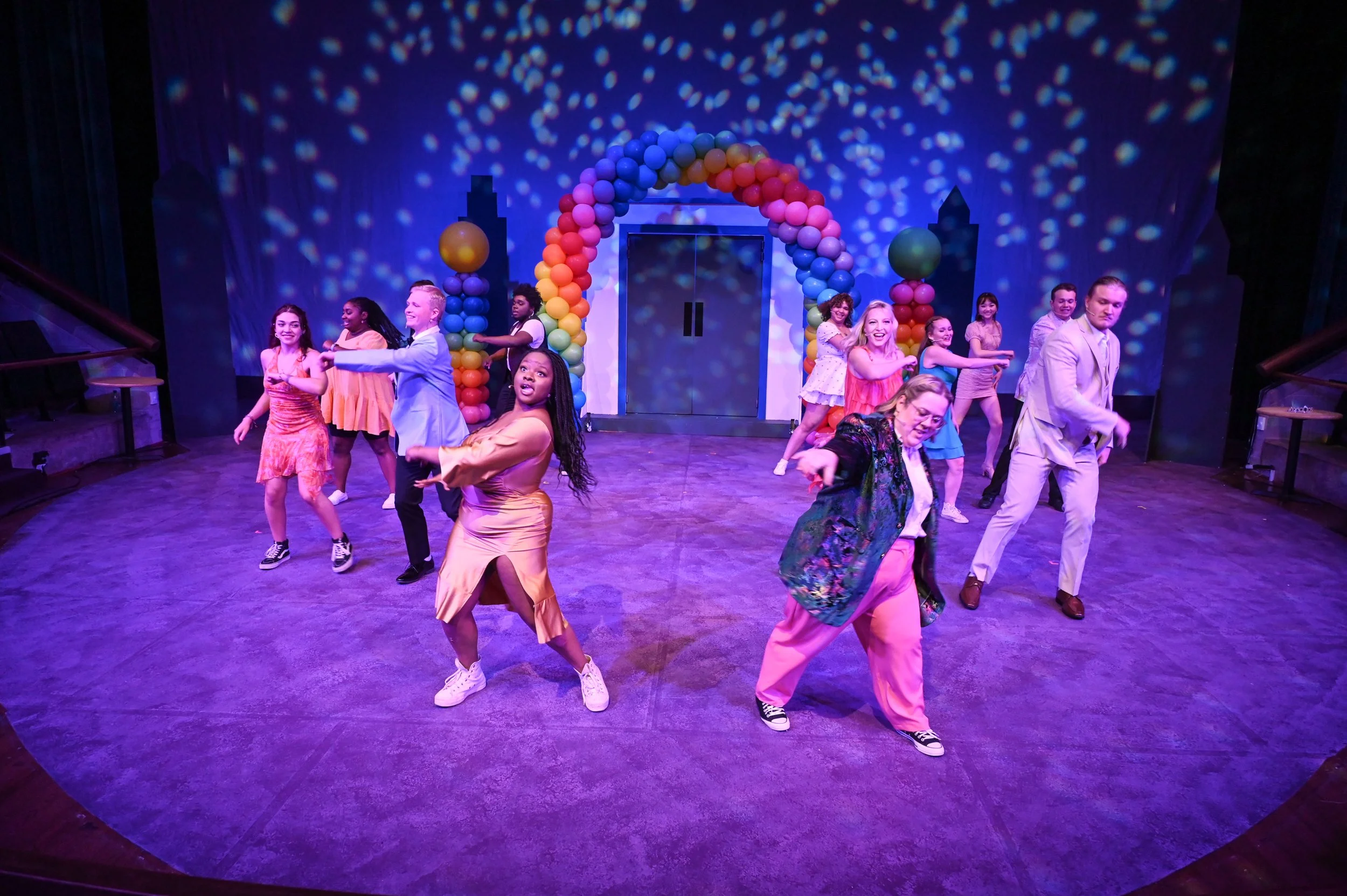 Group of people dancing on stage with colorful balloon arch and cityscape backdrop at a lively performance.